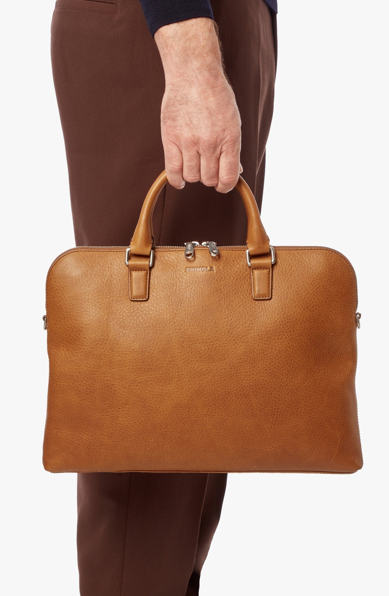 A person in brown pants holds the Shinola Portfolio Briefcase – Tan, featuring double handles and silver hardware, against a plain white background.