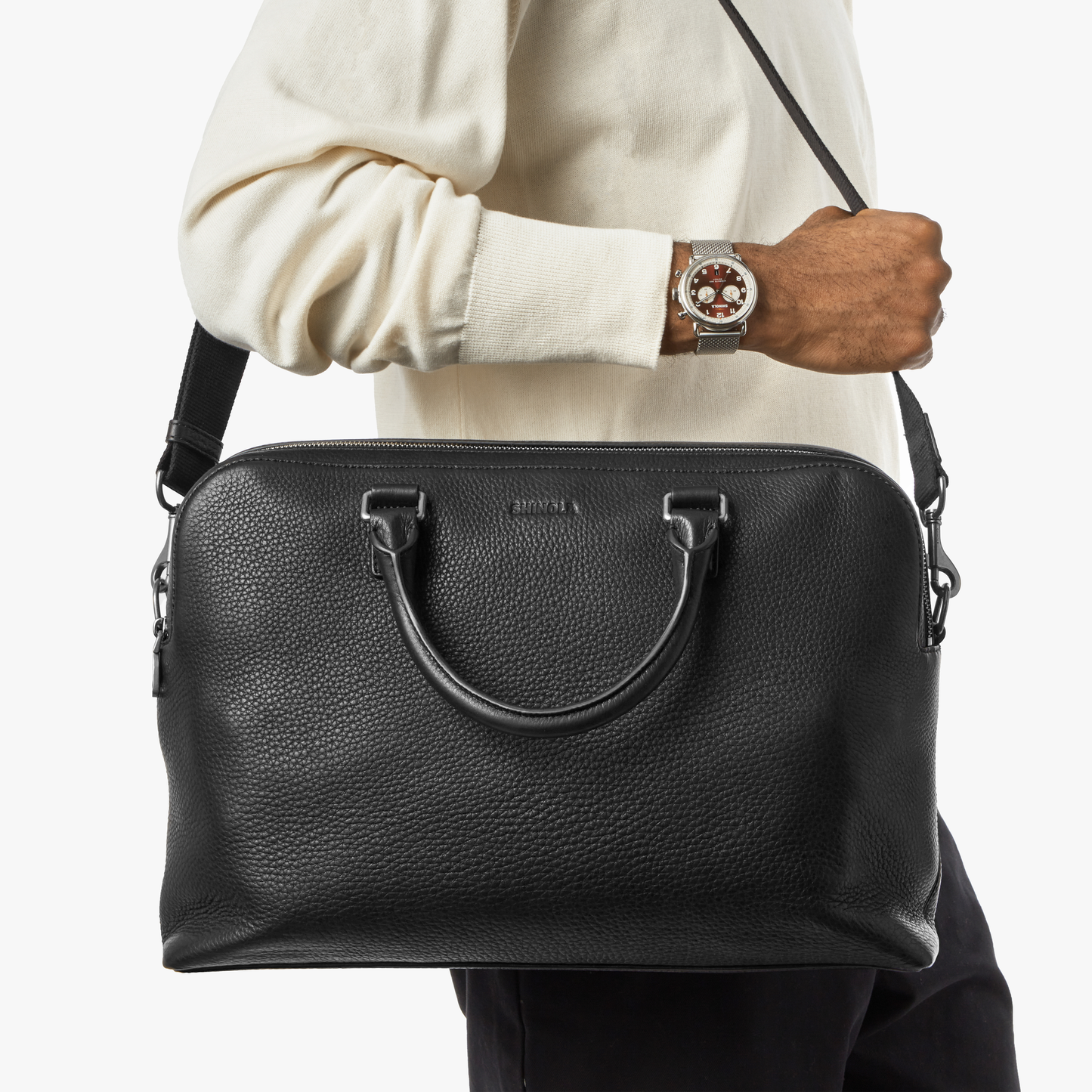 A person in a cream sweater and watch holds the Shinola Double Zip Briefcase - Black, made of Natural Grain leather with both handle and shoulder strap, plus an extra compartment, against a plain white background.