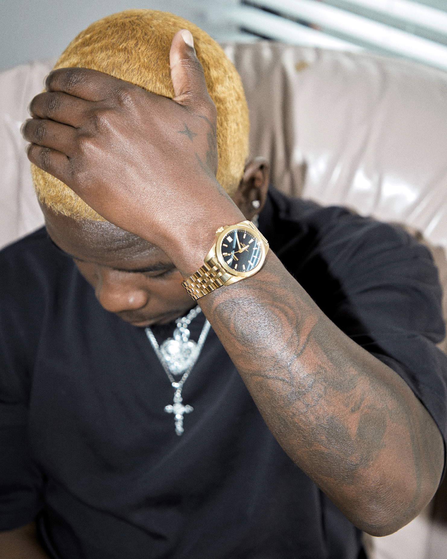 A person with bleached blonde hair, tattoos on their forearm, and a gold Shinola Circadian Monster Automatic Watch runs their hand over their head. They wear a black shirt, silver chain necklace, and sit on a light sofa.