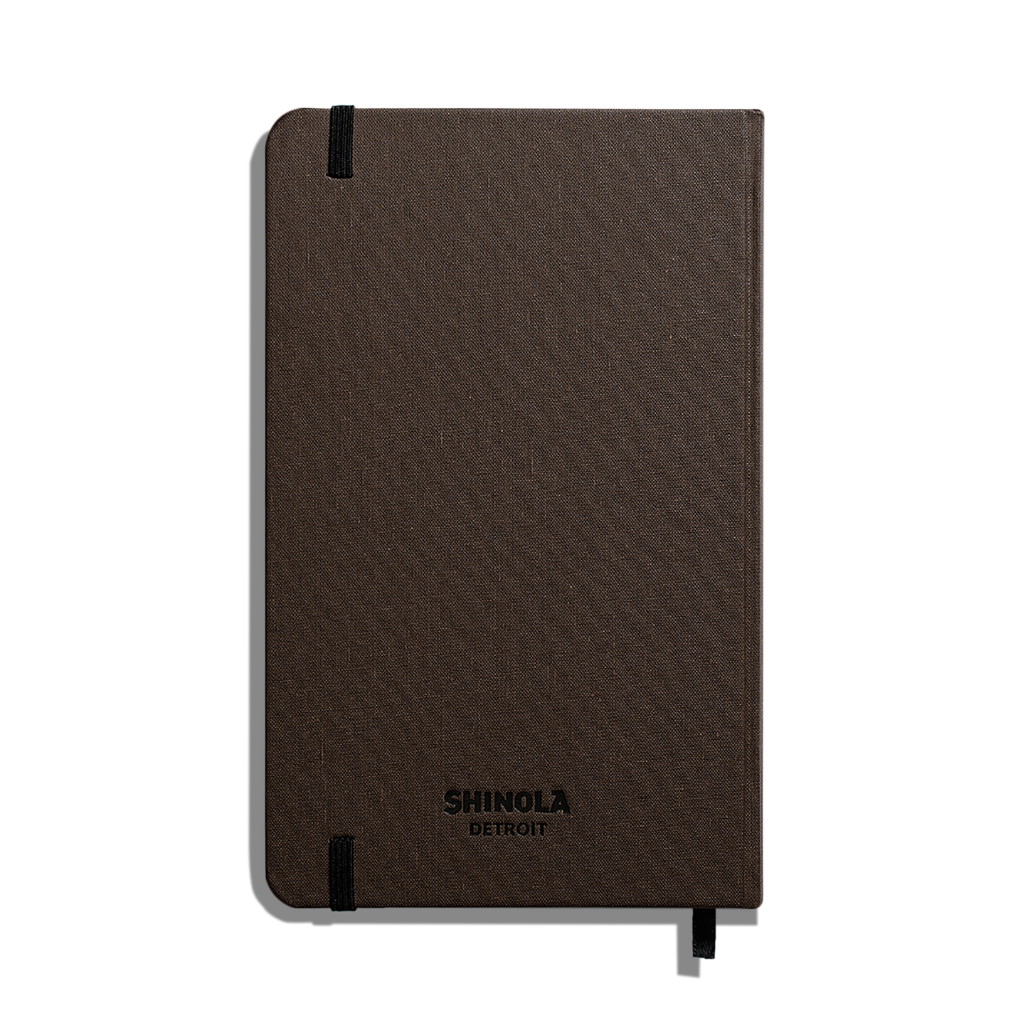 A closed, dark brown Shinola Medium Hard Ruled Journal (5.25" x 8.25") features a textured cover, Smyth-sewn binding, black elastic band on the top left, and a ribbon bookmark at the bottom right.