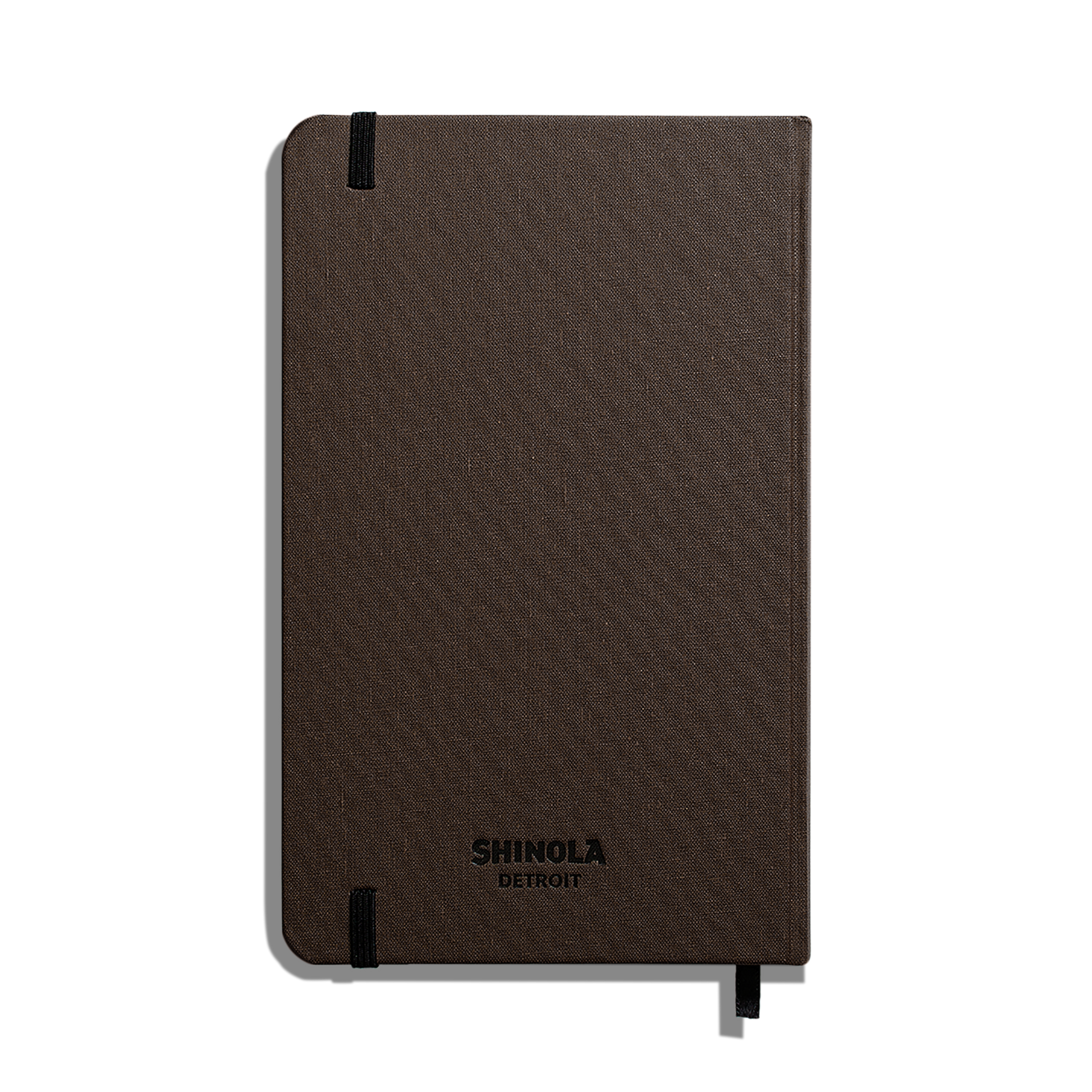 A closed, dark brown Shinola Medium Hard Ruled Journal (5.25" x 8.25") features a textured cover, Smyth-sewn binding, black elastic band on the top left, and a ribbon bookmark at the bottom right.