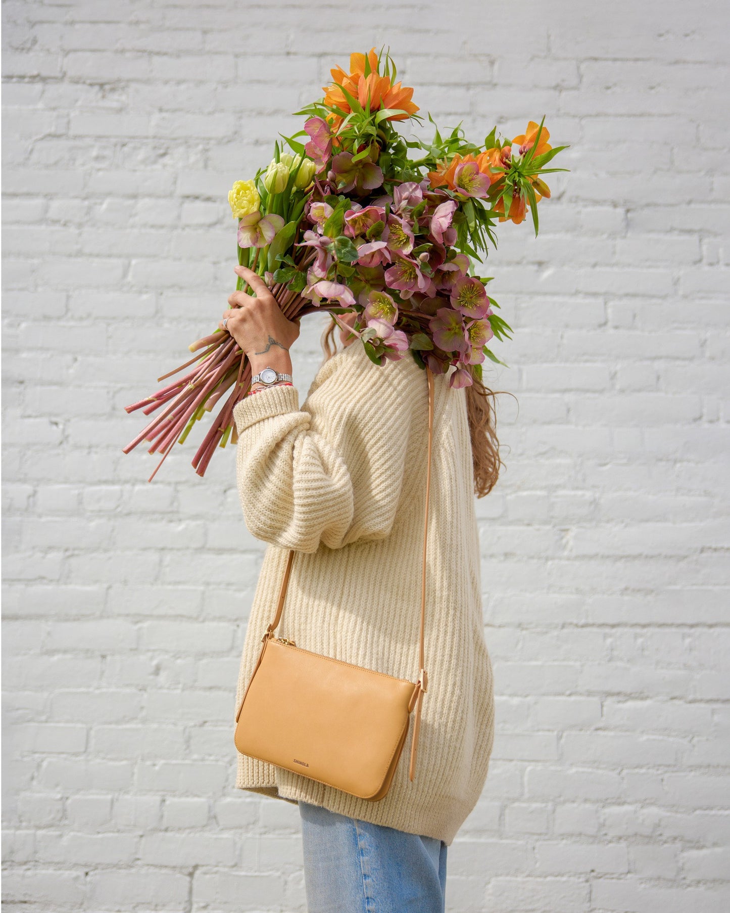 A person in a cream sweater and jeans stands by a white brick wall, holding a bouquet in front of their face. They wear a watch and carry the Shinola Small Bixby Crossbody - Honey, an everyday bag made from natural leather.