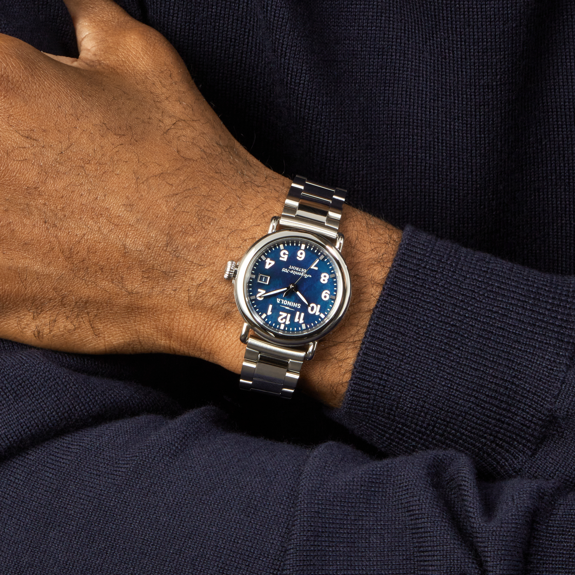 A person in a dark blue sweater stands with crossed arms, displaying a Shinola Runwell Watch - Cadet Blue with a mother of pearl dial, white numerals, and a stainless-steel bracelet on their left wrist.