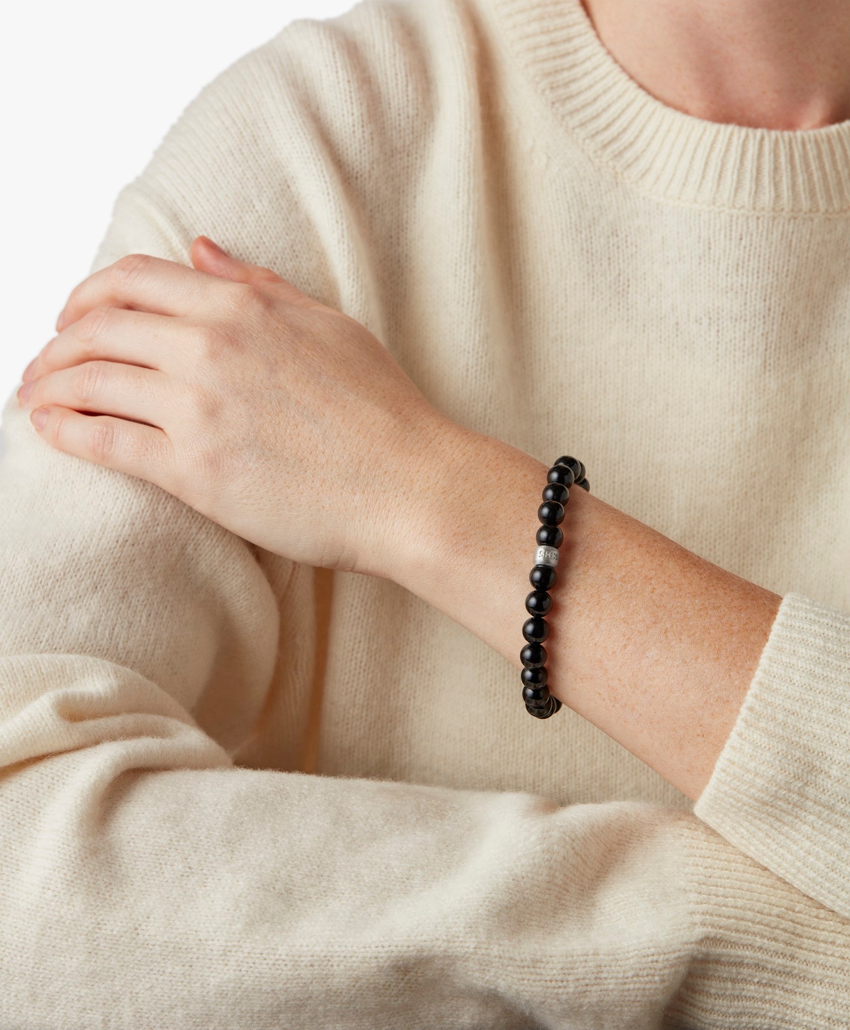 A person in a cream sweater rests an arm across their chest, showcasing the Shinola 8mm Beaded Onyx Bracelet – Onyx with a sterling silver accent on their wrist.