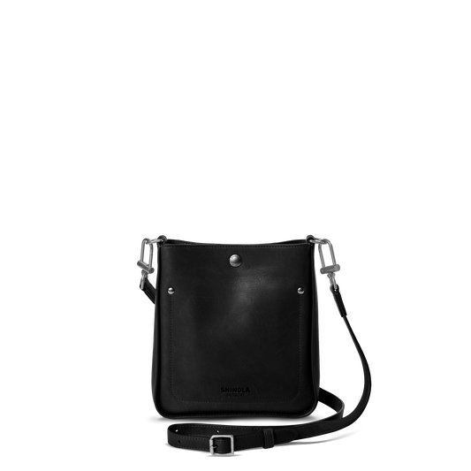The Shinola Mini Pocket Crossbody Bag - Black features silver hardware, a front snap button, and an adjustable leather strap—ideal for carrying everyday essentials in style. Shown here against a plain white background.