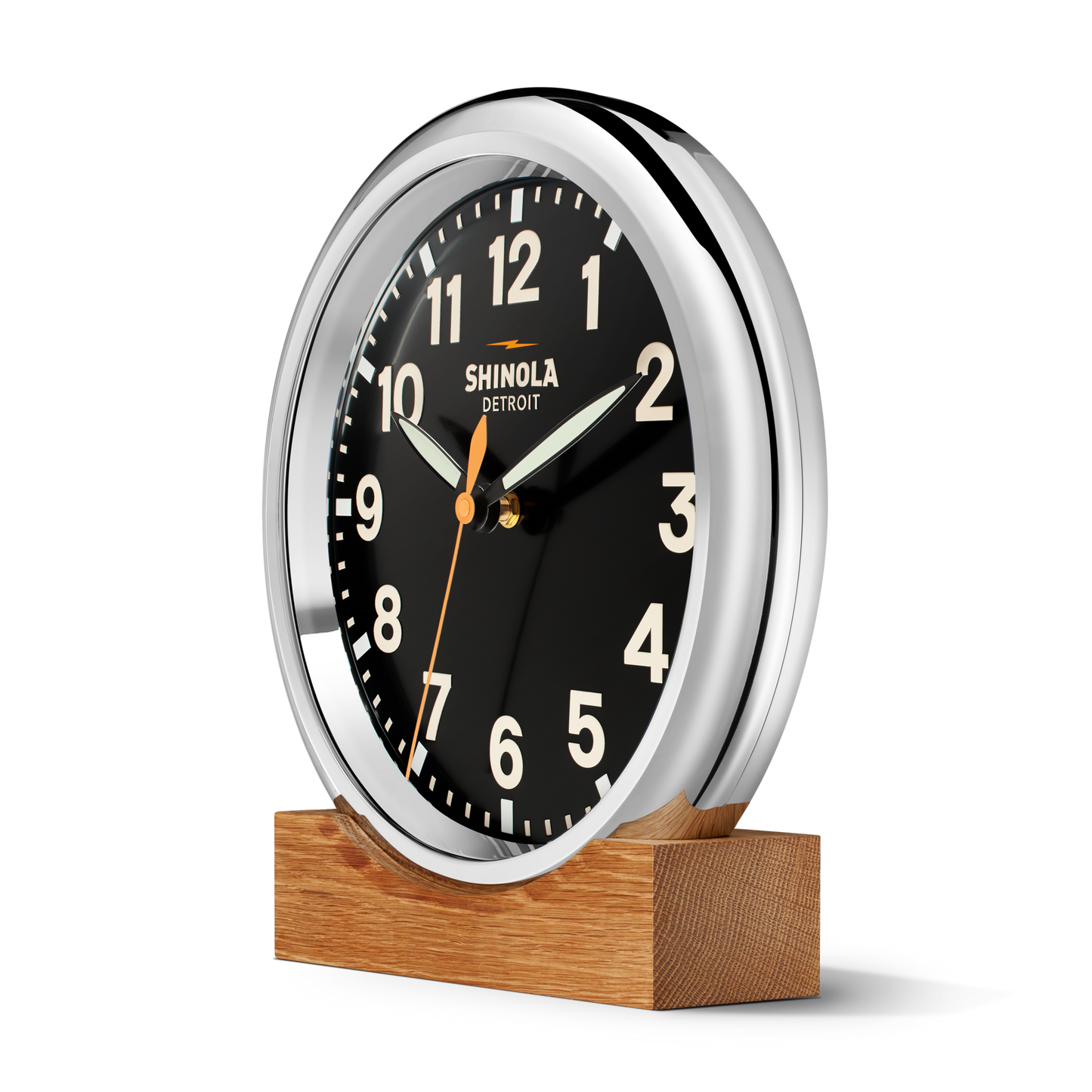 The 14" Runwell Clock by Shinola features a black face, white numbers, and chrome casing. Branded with Shinola Detroit, it sits on a rectangular wooden base and shows the time as 10:09:35.