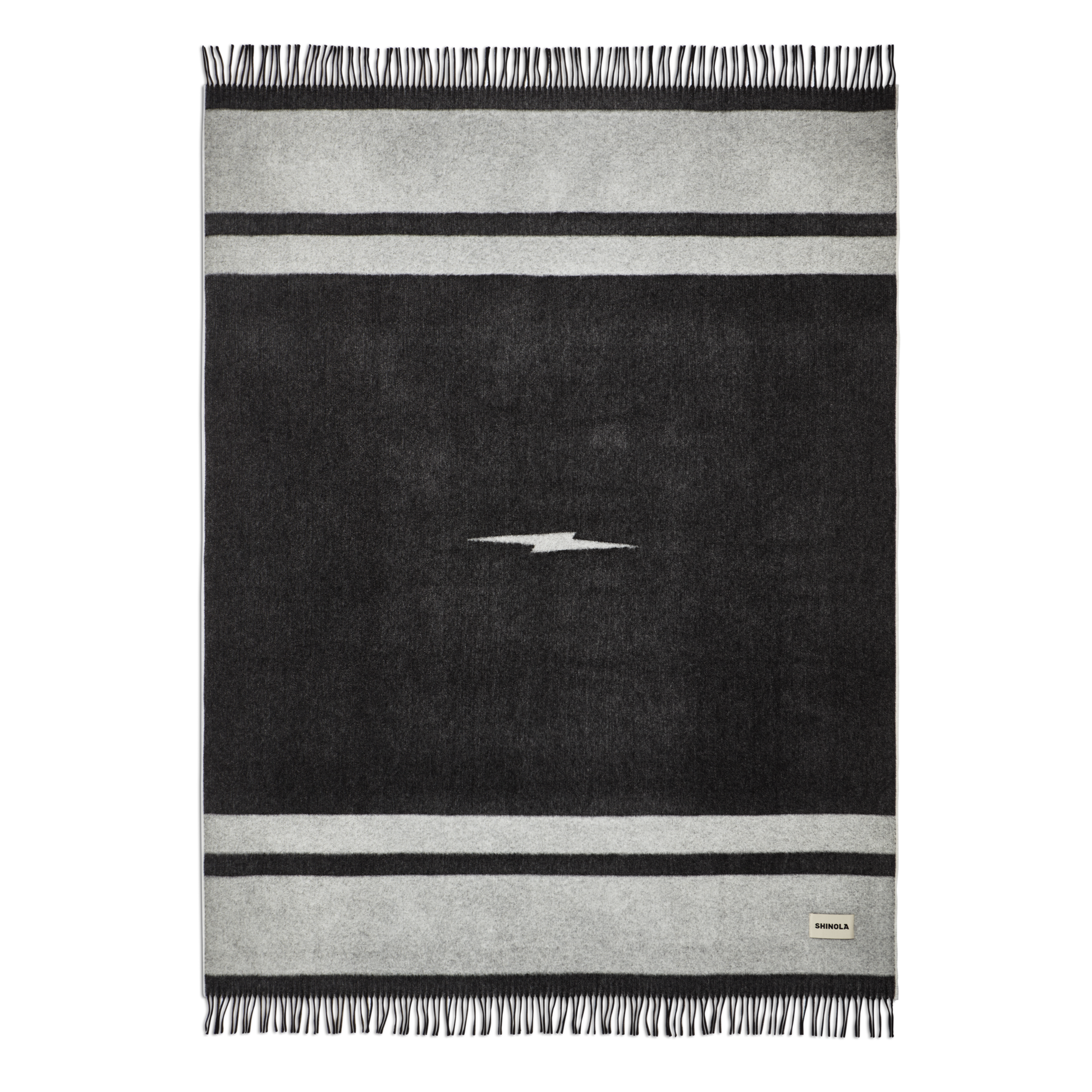 The Shinola Woven Alpaca Wool Blanket is a handcrafted black and gray rectangular throw from Peru, featuring a central white lightning bolt, two horizontal gray stripes, fringe on the short edges, and a small corner fabric label.