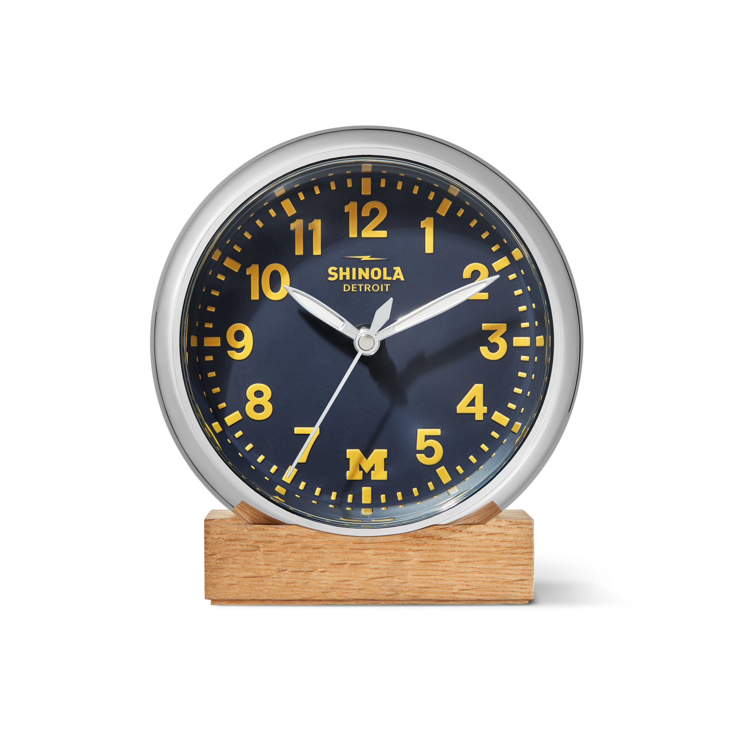 The 6" Runwell Clock - Blue by Shinola is a round analog desk clock with a wolverine blue face, yellow numbers and hands, the letter M at the bottom, Shinola branding at the top, reads 2:10, and sits on a wooden base.