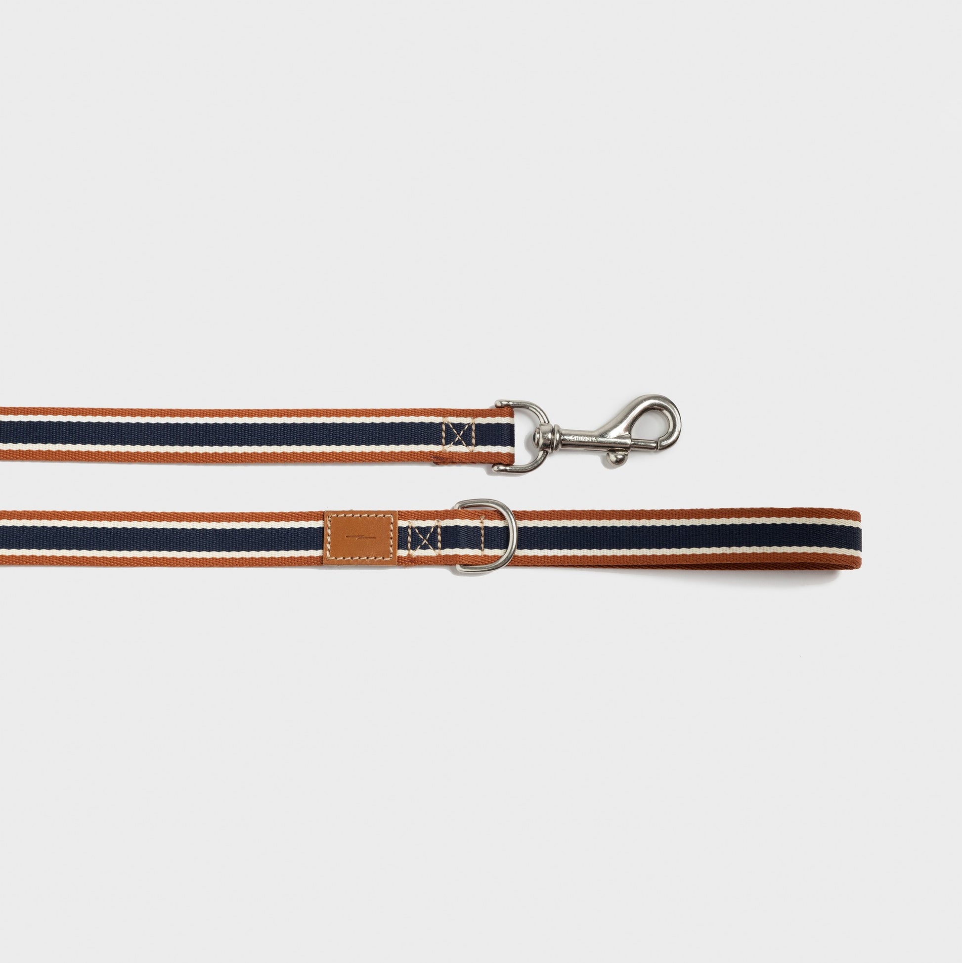 The Shinola Nylon Dog Leash - Brand Stripe features durable nylon in orange, navy blue, and white stripes, a silver metal clasp, leather patch, and metal D-ring. The leash is shown laid flat on a white background.