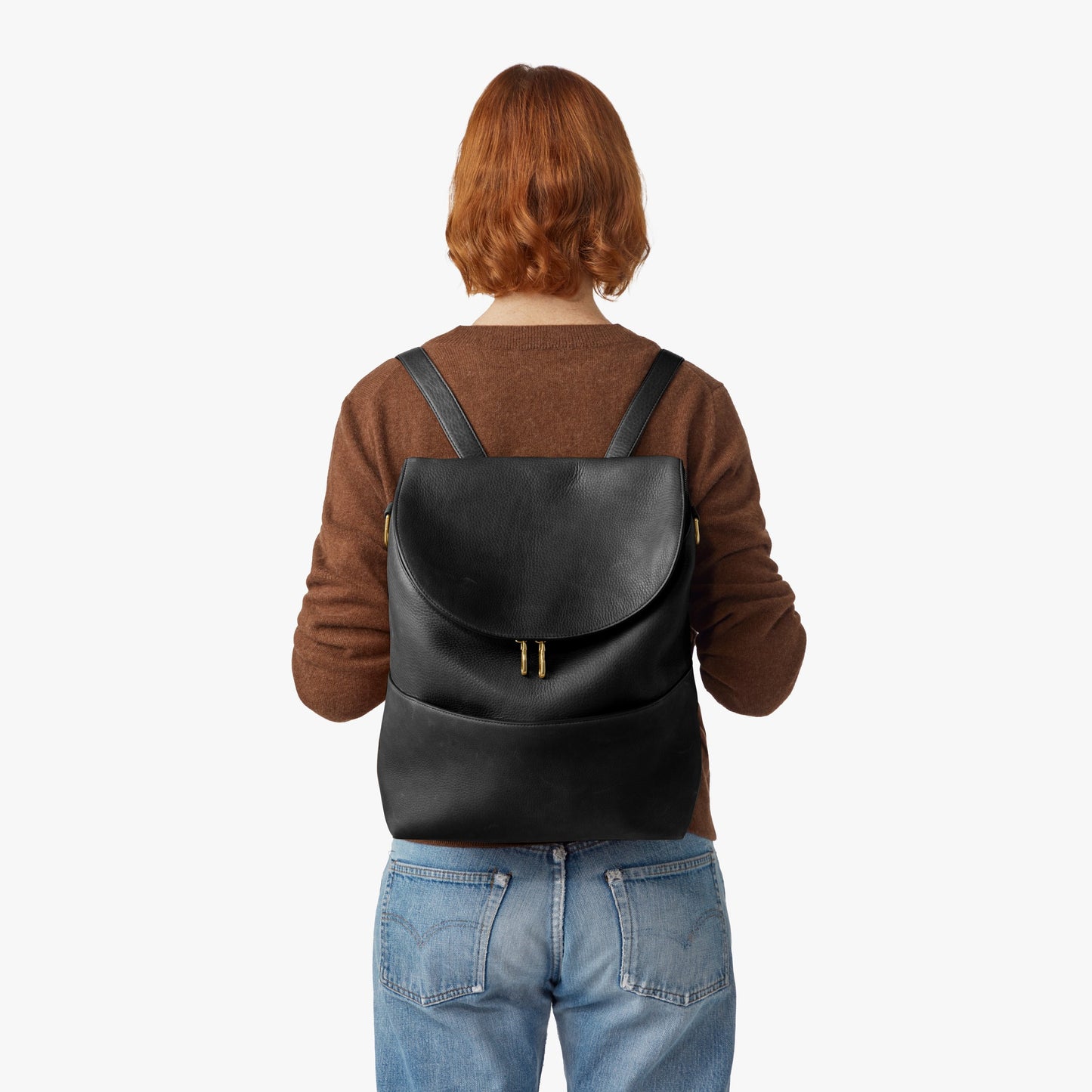 A person with short red hair, in a brown sweater and blue jeans, is seen from behind carrying the Shinola Convertible Pocket Backpack - Brand Stripe / Black against a plain white background.