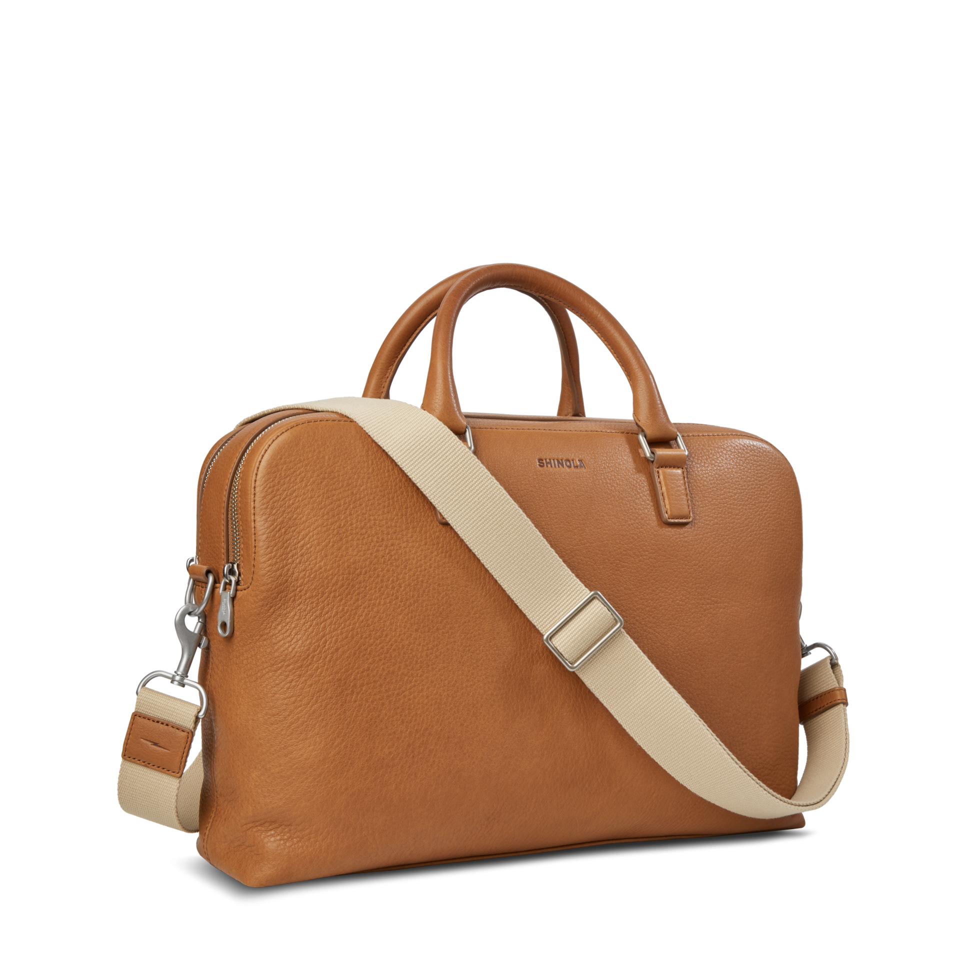 The Shinola Double Zip Briefcase - Tan in Natural Grain leather features two short handles, an extra compartment, a beige adjustable detachable shoulder strap, zippered top, and silver hardware, shown against a plain white background.