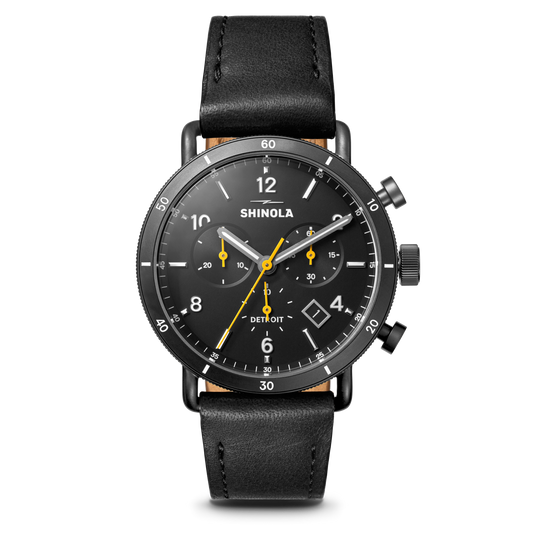 The Shinola Canfield Sport Chronograph Watch features a black leather strap, gunmetal stainless steel case, black dial with white numerals and yellow hands, plus subdials and the brand name on its face.