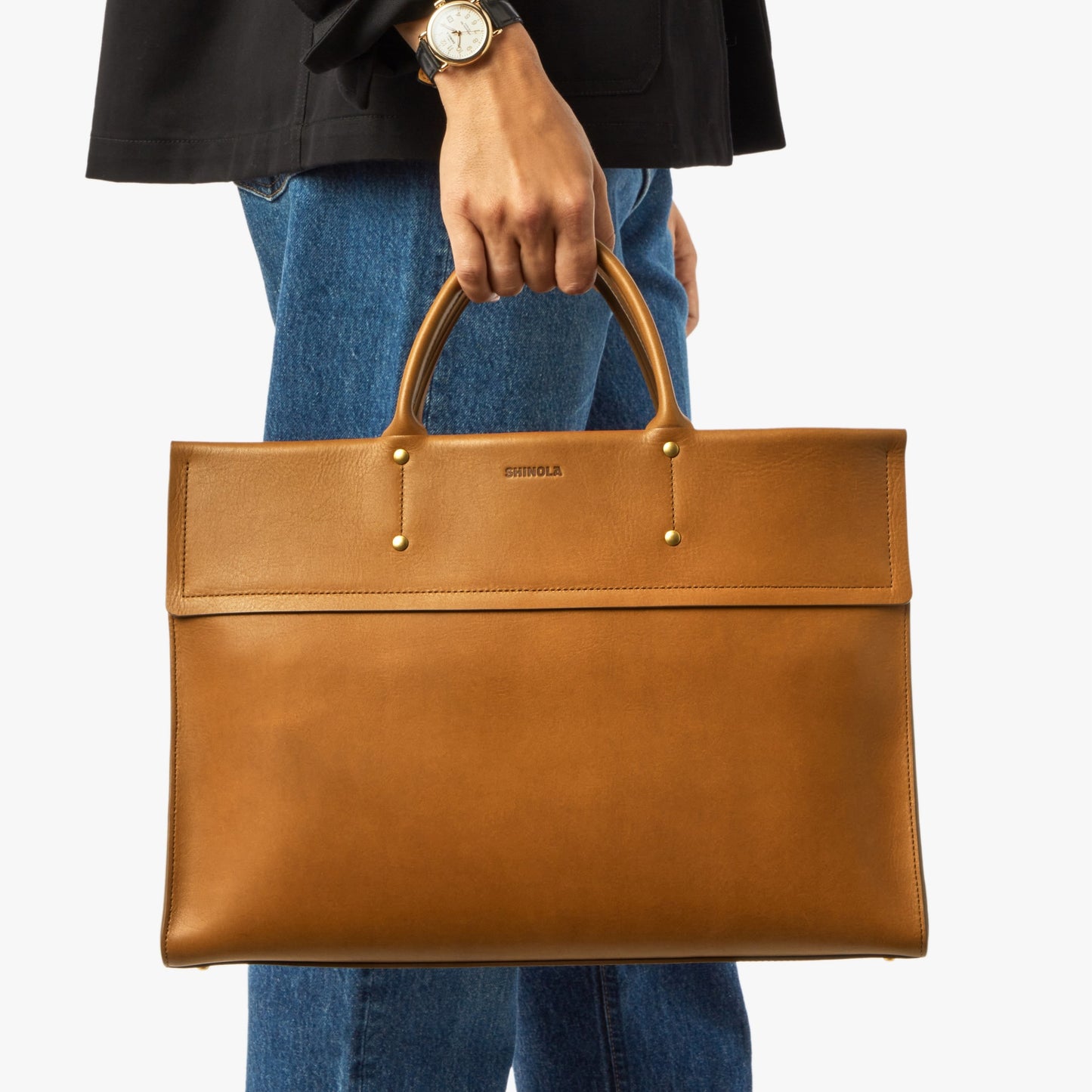 A person in blue jeans and a black jacket holds the Shinola Day Satchel - Tan by its handles. Only their lower torso and hand are visible. The satchel features gold hardware, "Shinola" on the front, and a spacious laptop compartment.