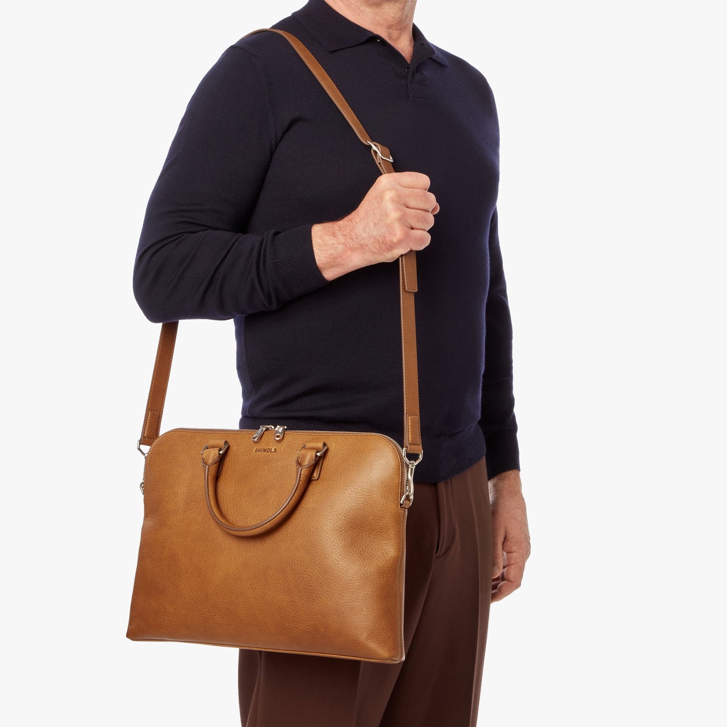 A man in a dark long-sleeve shirt and brown pants carries the Shinola Portfolio Briefcase - Tan, featuring pebbled leather, handles, and a shoulder strap, against a plain white background.
