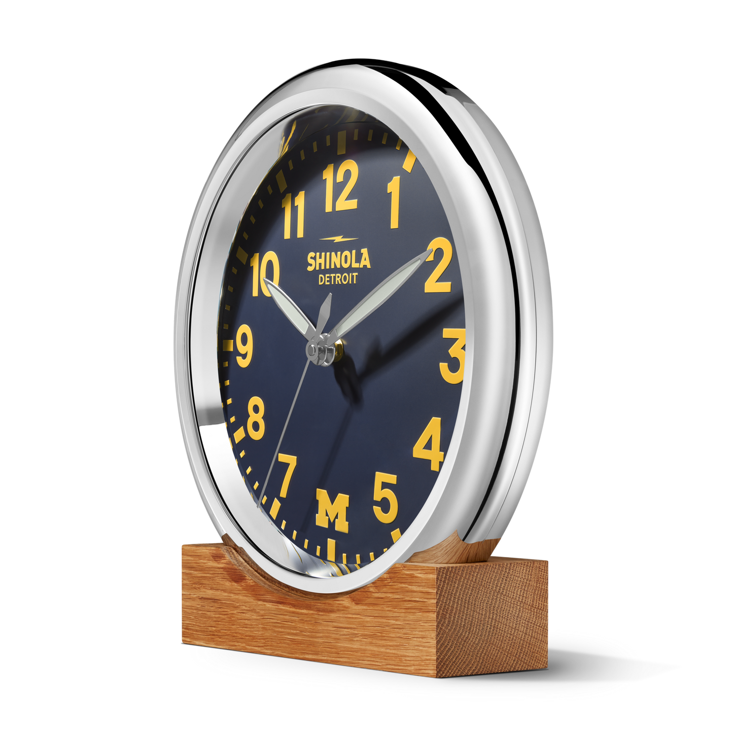 The 14" Runwell Clock - Blue by Shinola features a navy blue face, yellow numbers, silver trim, and an M logo nodding to the University of Michigan, with SHINOLA DETROIT on the dial. It sits elegantly on a wooden stand.