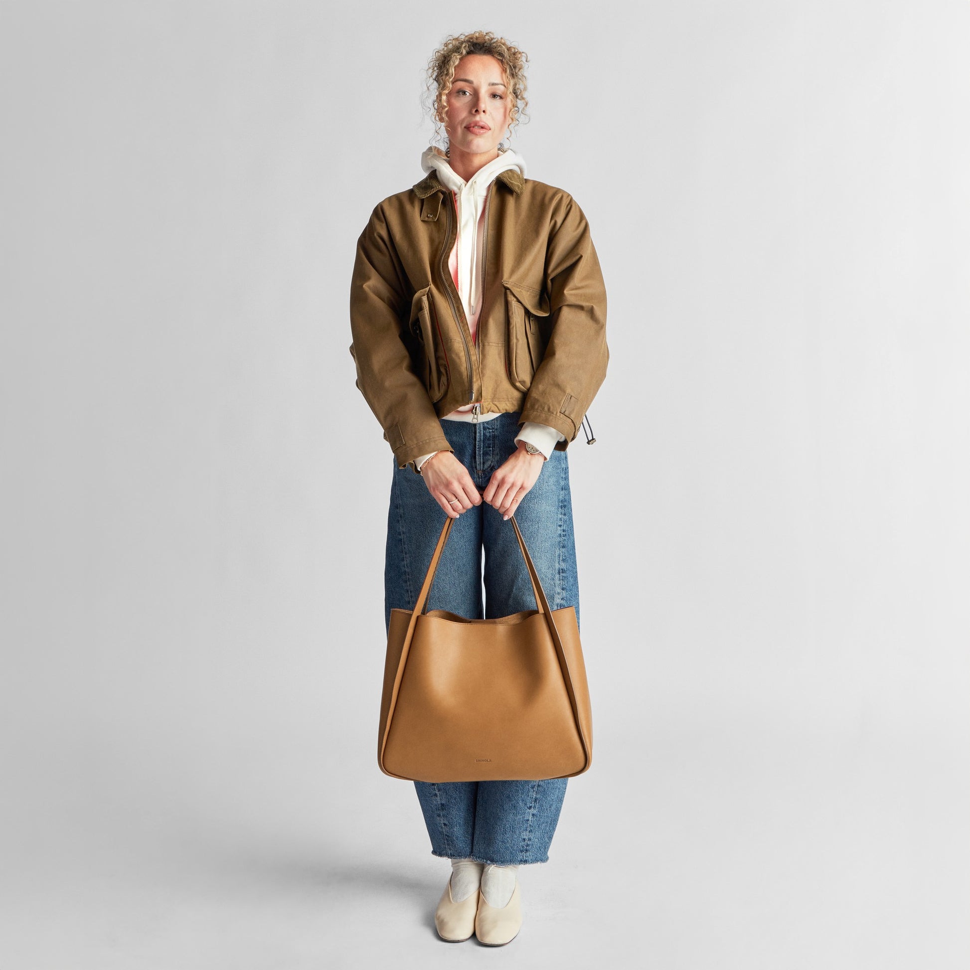 A person stands against a plain background, wearing a brown jacket, white hoodie, blue jeans, and white shoes, holding the Shinola Derby Tote Bag - Tan in front of them with both hands—an ideal choice for everyday use.