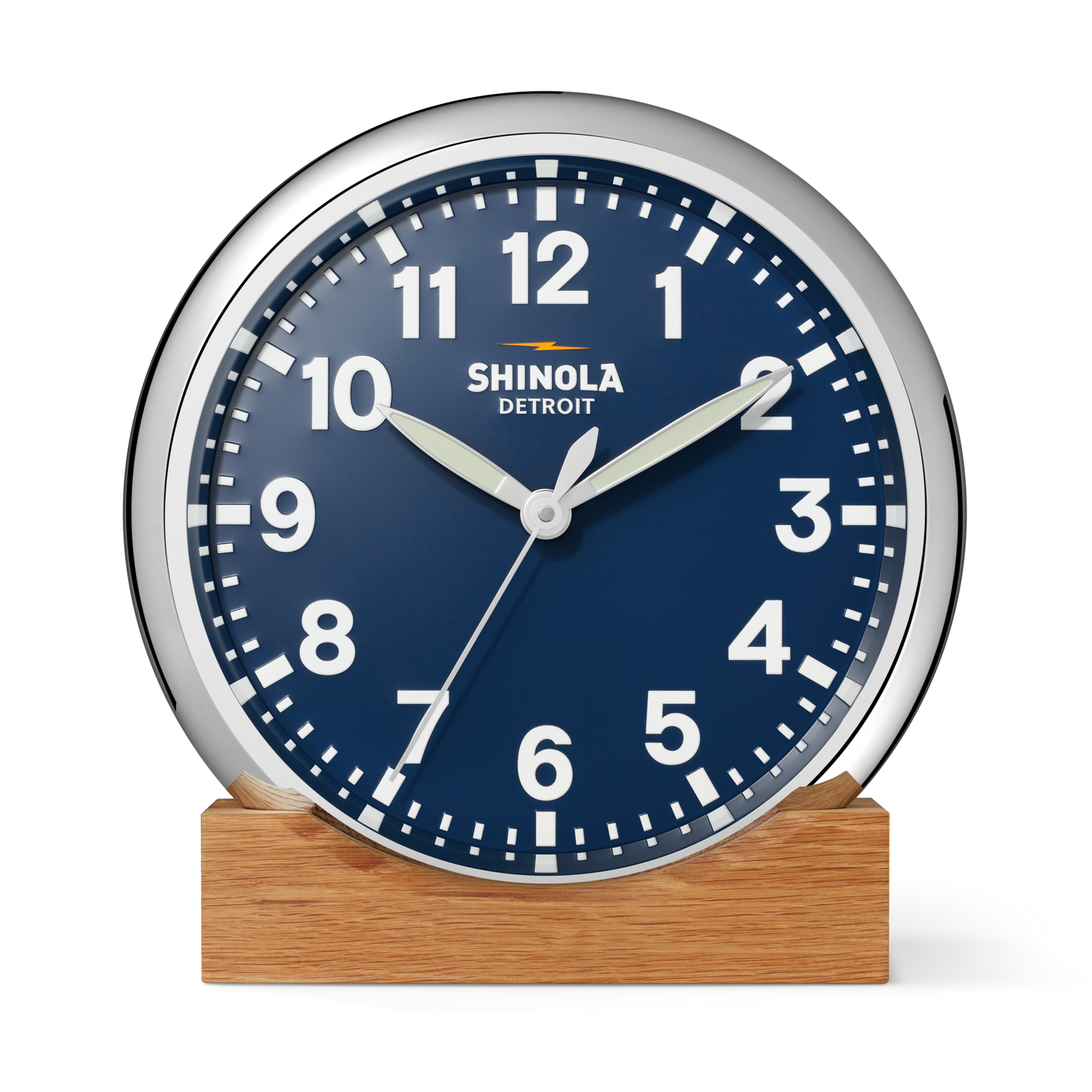 A Shinola 14" Runwell Clock - Navy with a matte midnight blue dial, white numbers, and silver frame sits on a wooden base. The clock reads 1:50:35, with "Shinola" written below the 12 o’clock mark.