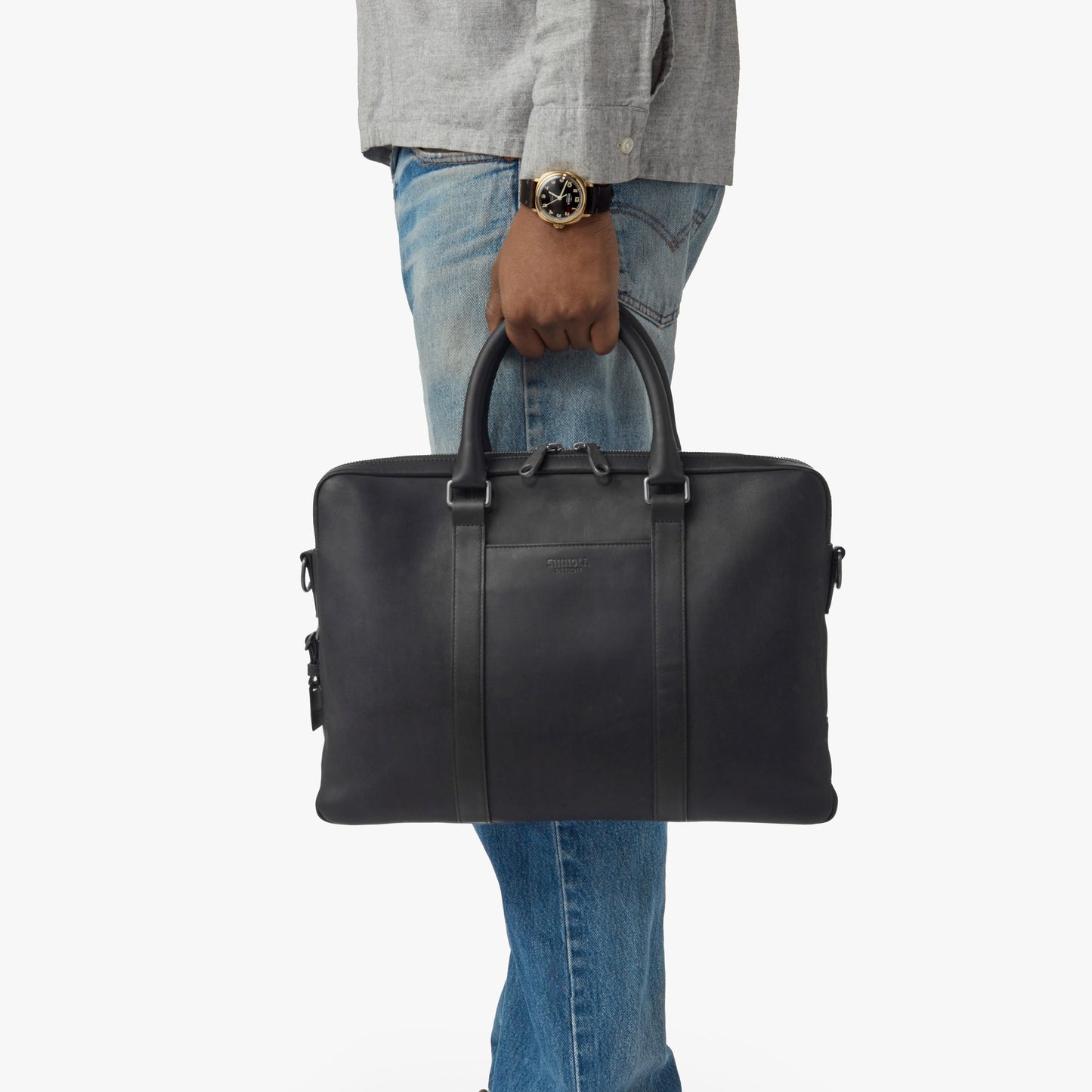 A person wearing a gray shirt, blue jeans, and a watch holds the shinola.momputer Briefcase - Black by its handles against a plain white background.