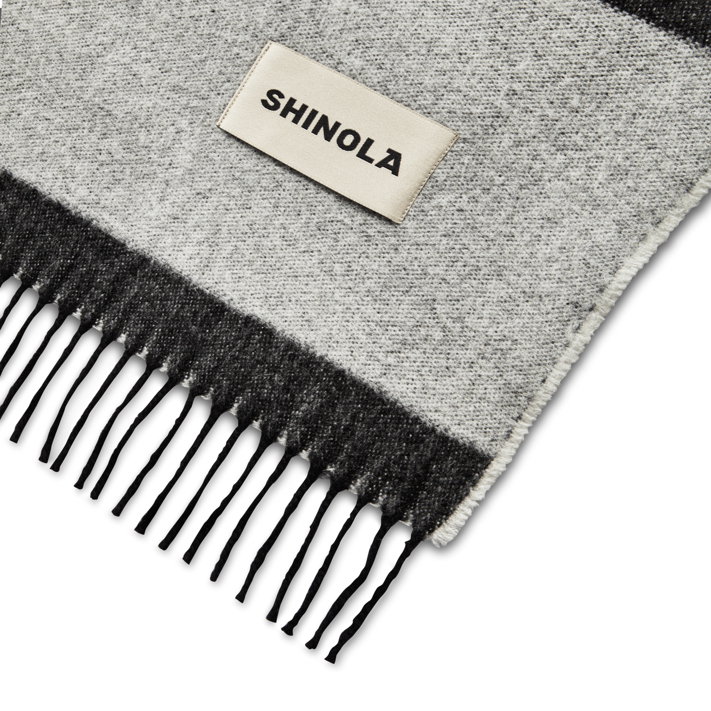A close-up of the Shinola Woven Alpaca Wool Blanket, featuring gray and black fringes and a rectangular white Shinola patch, reminiscent of premium Peruvian craftsmanship.