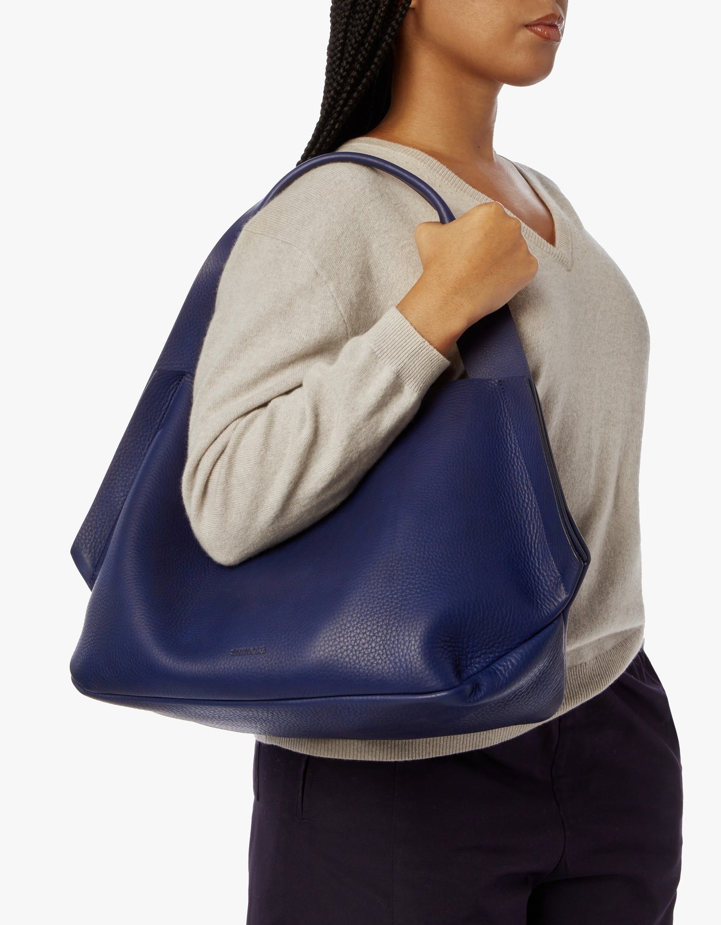 A person in a light beige sweater and dark pants carries the Shinola Runwell Shoulder Bag - Lake, featuring a textured surface, relaxed structure, and handy leather card pocket, on their left shoulder.