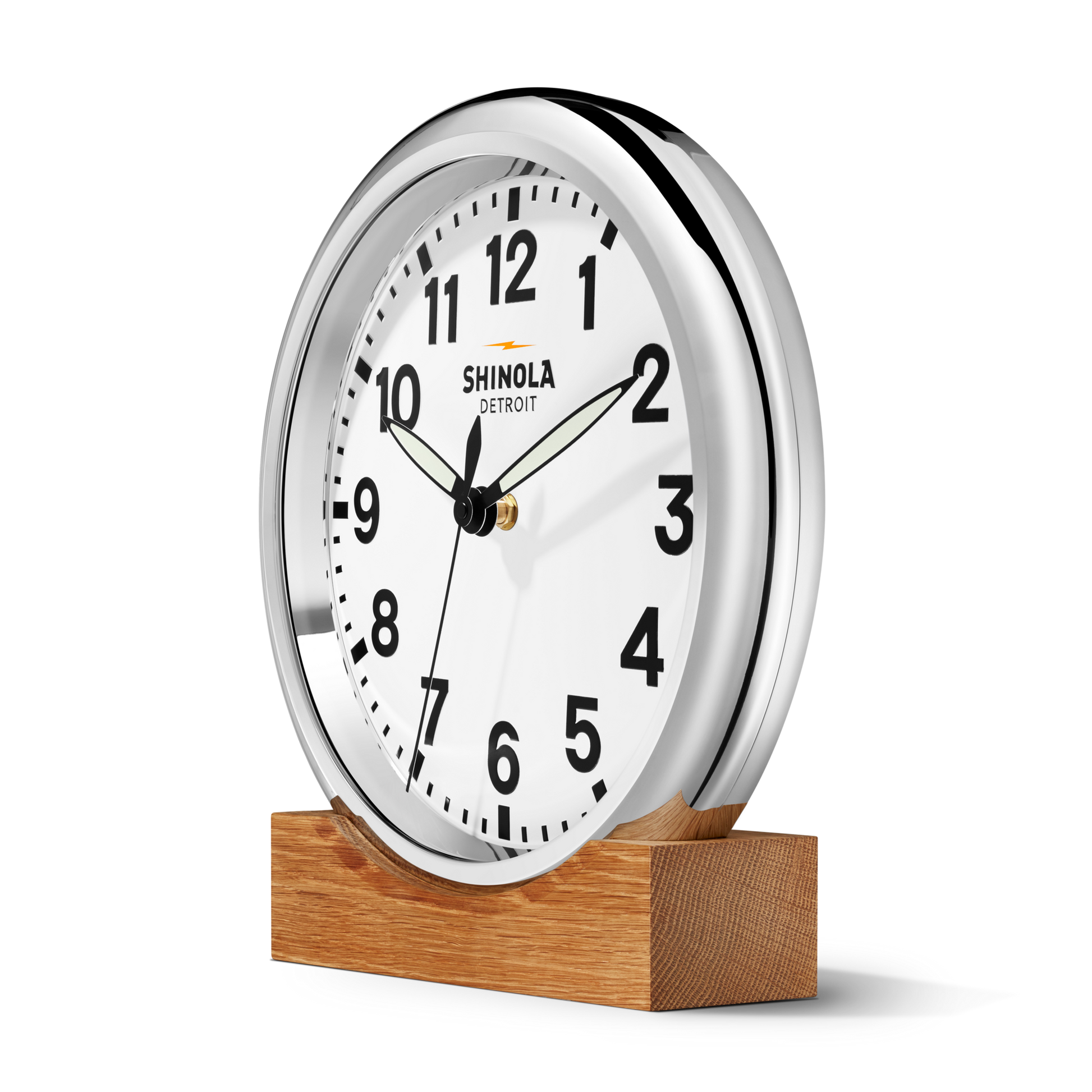 The Shinola 14" Runwell Clock features a chrome case, white face, black numbers, and hands. Set to 10:09, it rests on a wooden stand against a plain white background.