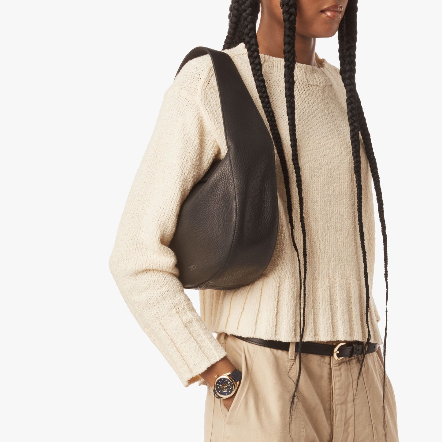 A person with long braids is seen from the lower face down, wearing a cream knit sweater and beige pants, holding the Shinola Small Shoulder Bag in Black natural grain leather.