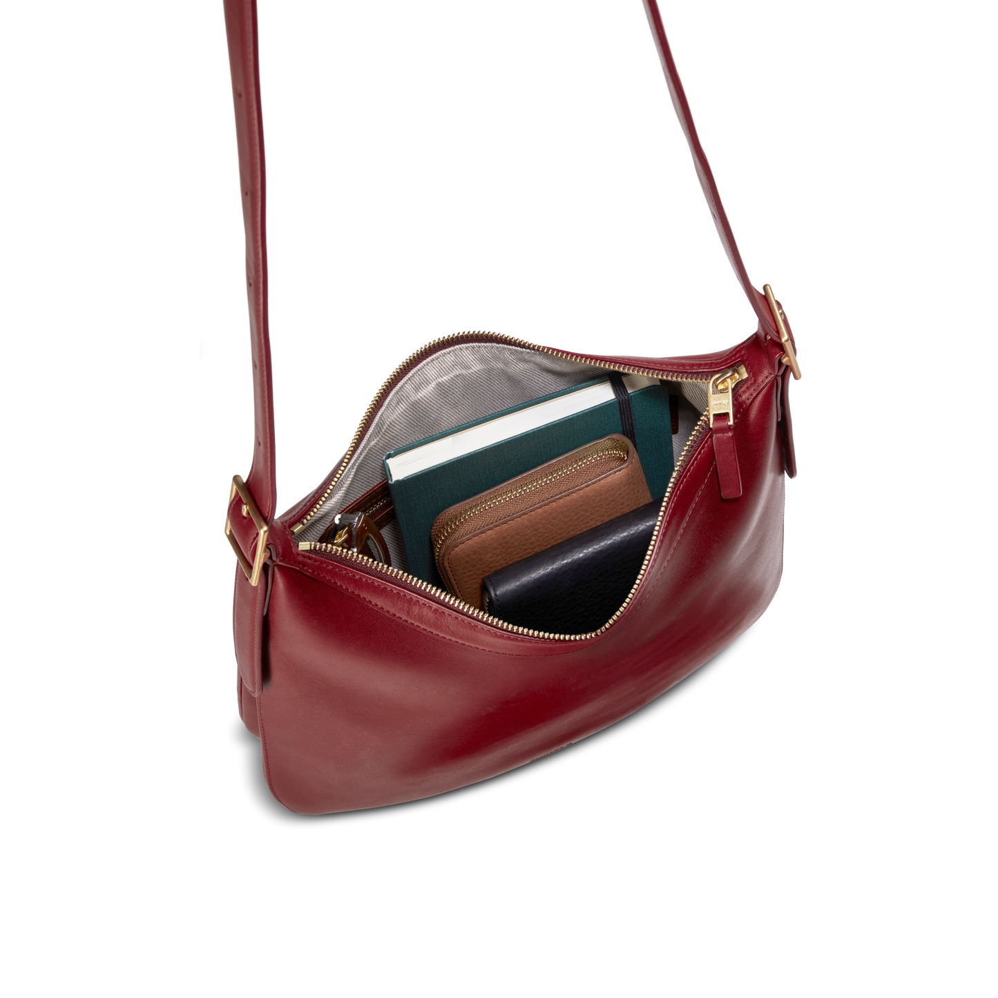 The Medium Bixby Crossbody - Ruby by Shinola features natural leather with a gold zipper and opens to reveal a green notebook, brown wallet, and black wallet—making it a versatile accessory for any occasion.