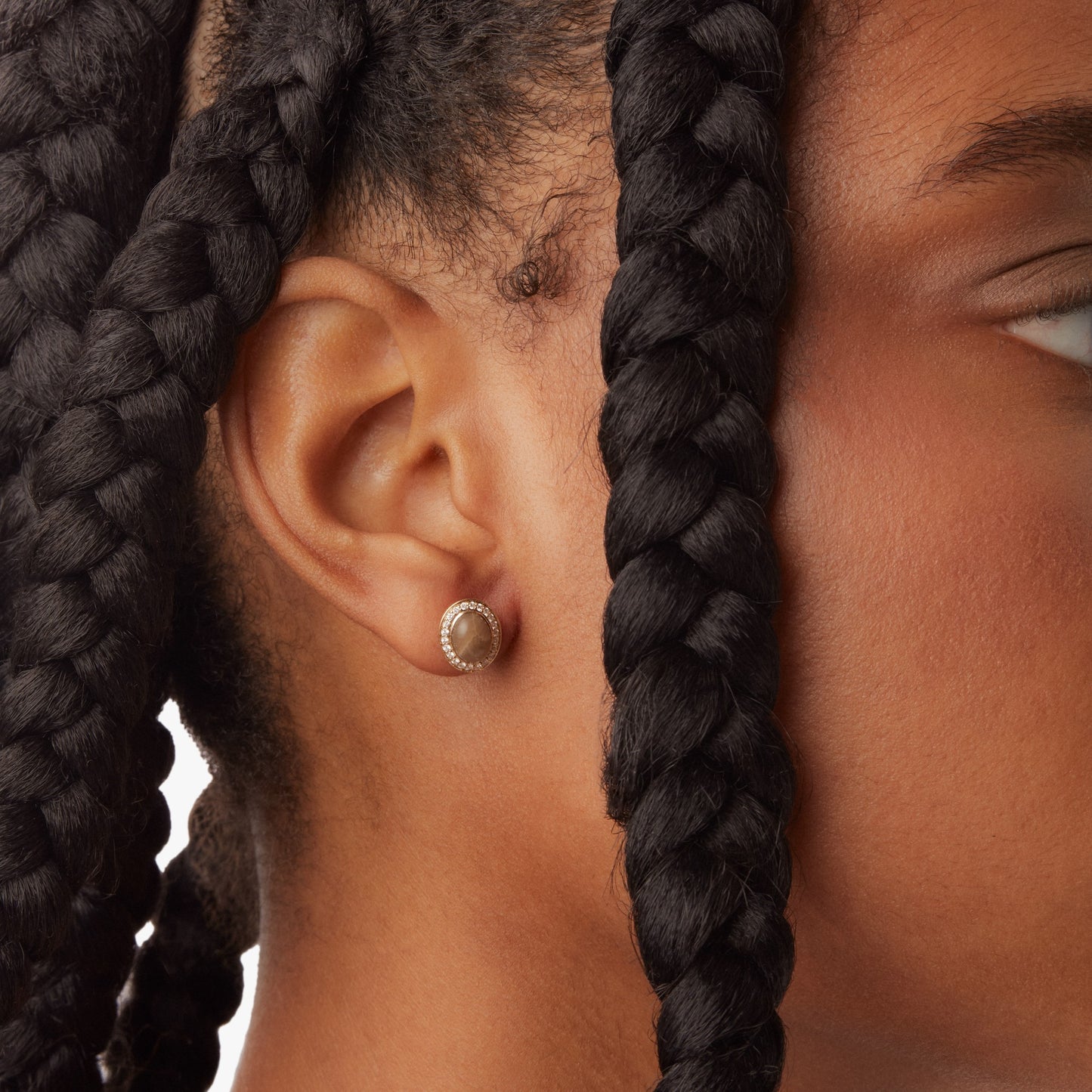 Close-up of a person’s ear with medium brown skin and thick, dark braids, wearing Shinola’s Diamond Pave Book Petoskey Studs in yellow gold. Photo is cropped to show only the ear and part of the face.