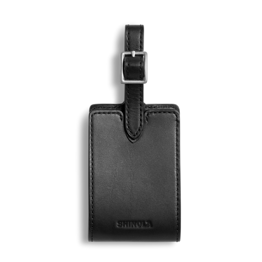 The Shinola Luggage Tag - Black, made from Italian Vachetta leather with a silver buckle and embossed branding, is shown on a white background. Personal monogramming is available.