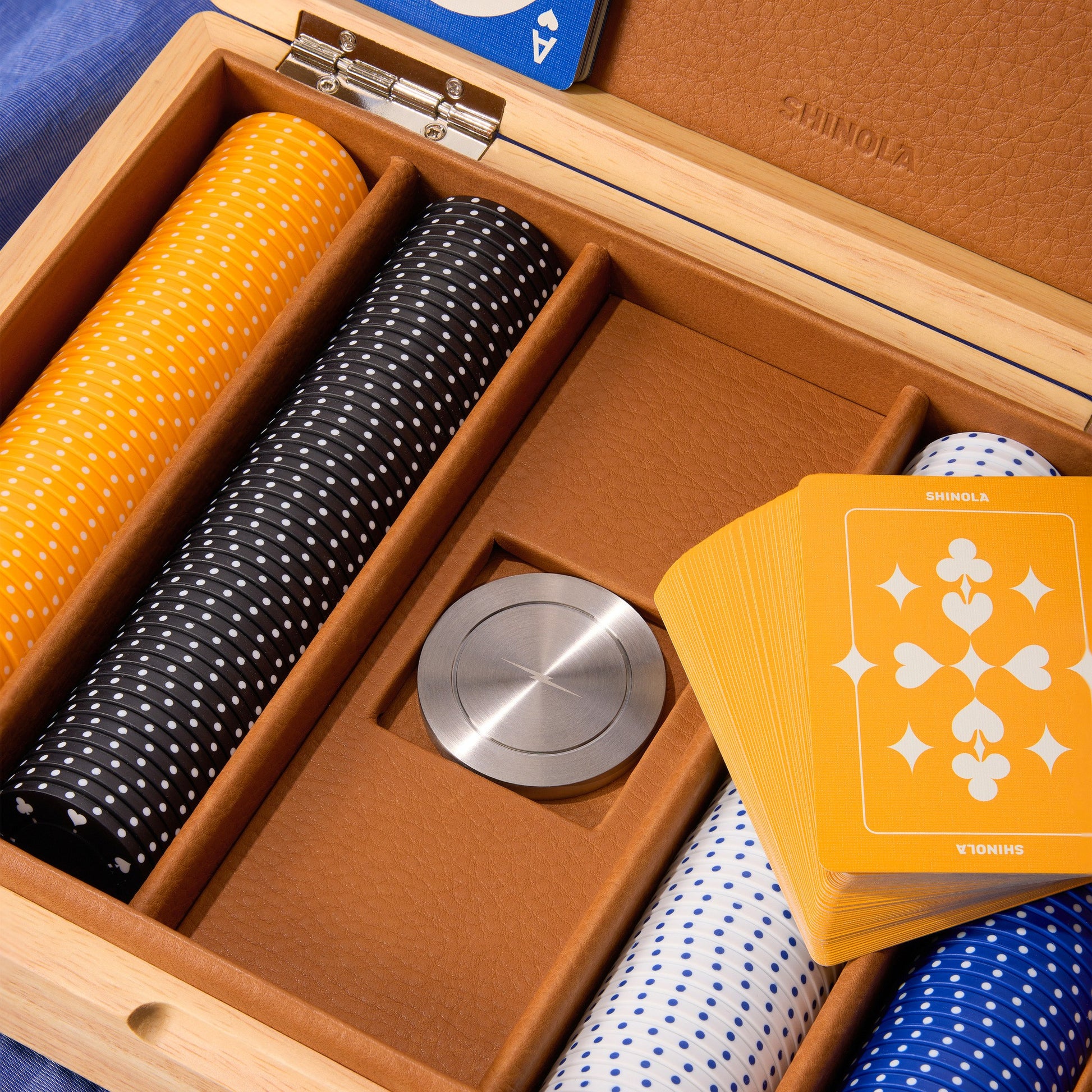 A close-up of the Shinola Lacquered Wood Poker Set in Cream/Blue, featuring custom poker chips, playing cards, and a metallic dealer button in a leather-lined, wood-accented case.
