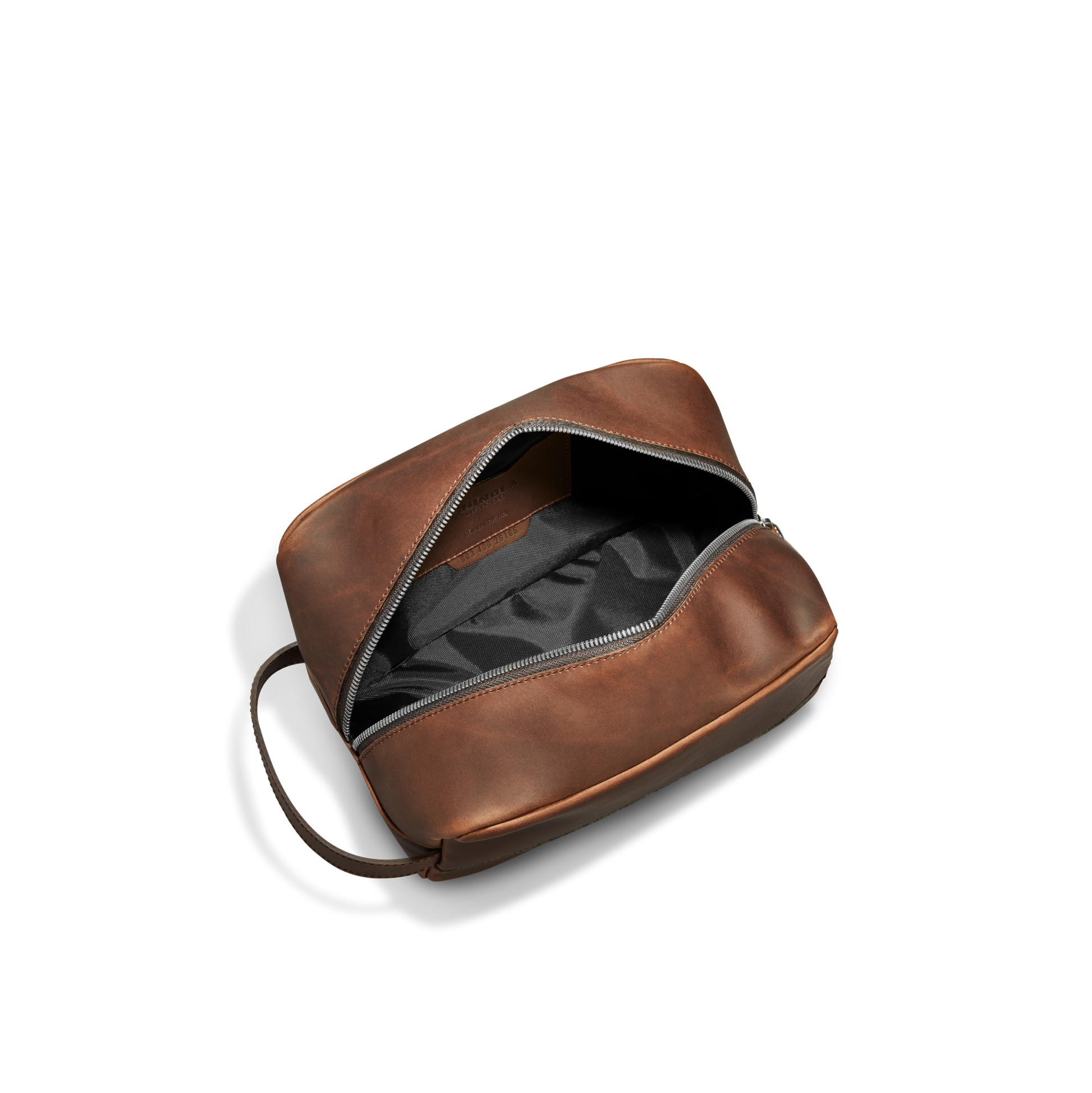 The Shinola Zip Travel Kit - Medium Brown is shown partially open, revealing a black water-resistant interior with compartments and a carrying handle, displayed on a white background.