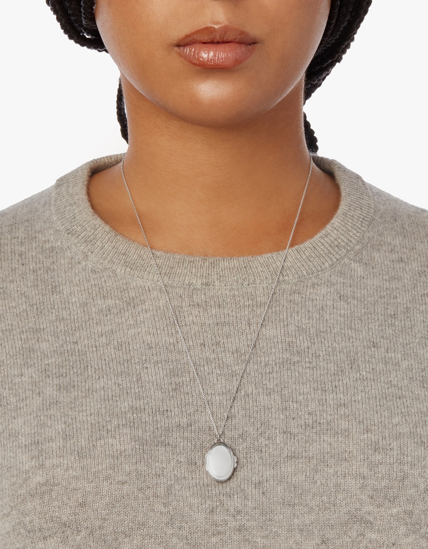 A woman wearing a light gray knit sweater and the Shinola Heirloom Statement Locket Necklace in sterling silver. The oval-shaped pendant is shown in a cropped shot from shoulders to just above the lips.