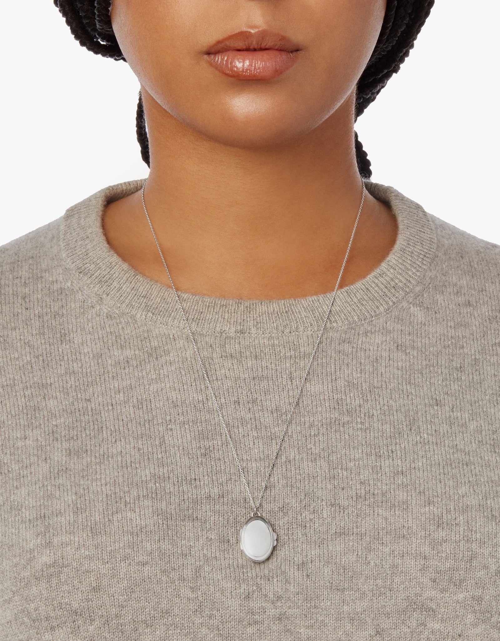 A woman wearing a light gray knit sweater and the Shinola Heirloom Statement Locket Necklace in sterling silver. The oval-shaped pendant is shown in a cropped shot from shoulders to just above the lips.