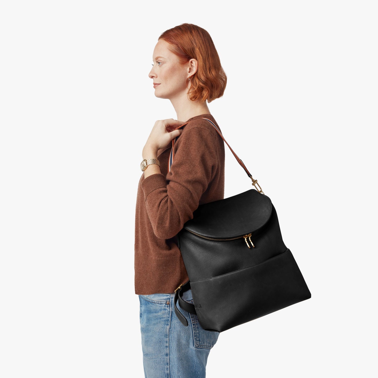 A person with short red hair, in a brown sweater and blue jeans, stands in profile holding the Shinola Convertible Pocket Backpack - Brand Stripe / Black over one shoulder against a plain white background.