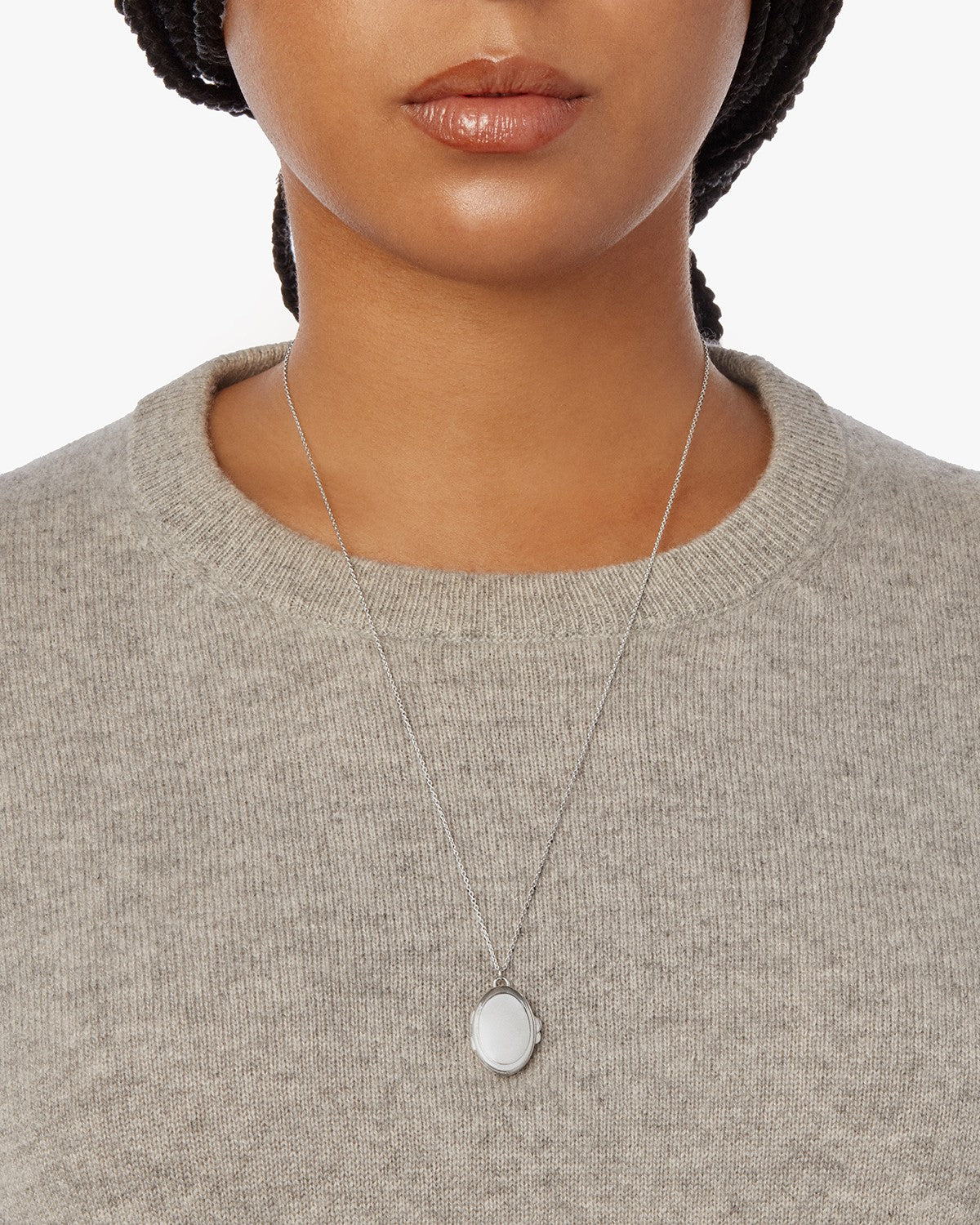 A person wearing a light gray knit sweater and the Shinola Heirloom Statement Locket Necklace in sterling silver, featuring an oval-shaped pendant, is shown from shoulders to chin against a plain white background.