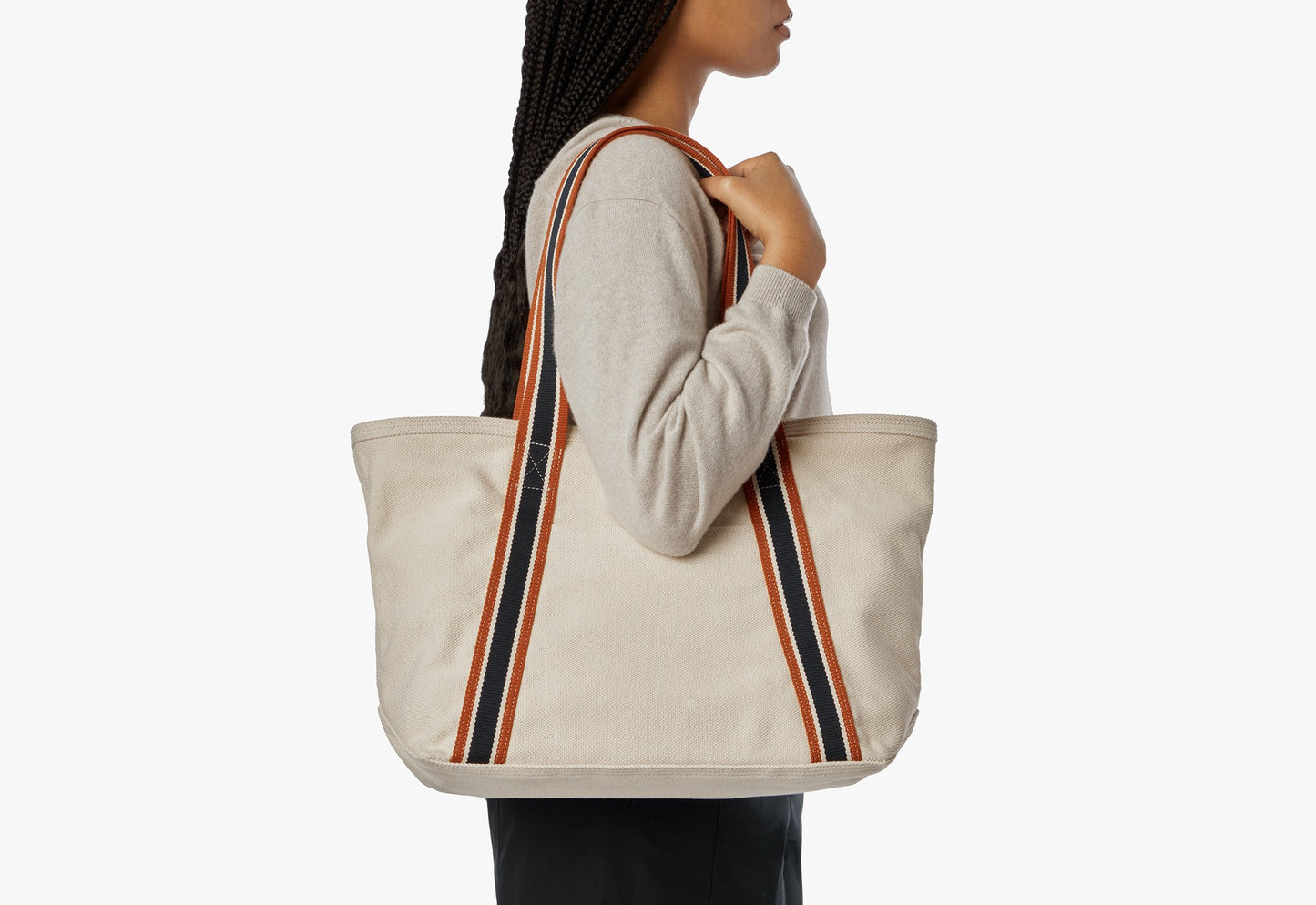A woman in a light sweater carries the Shinola Small Easy Canvas Tote - Natural, featuring black and orange striped handles, over her shoulder against a plain white background.