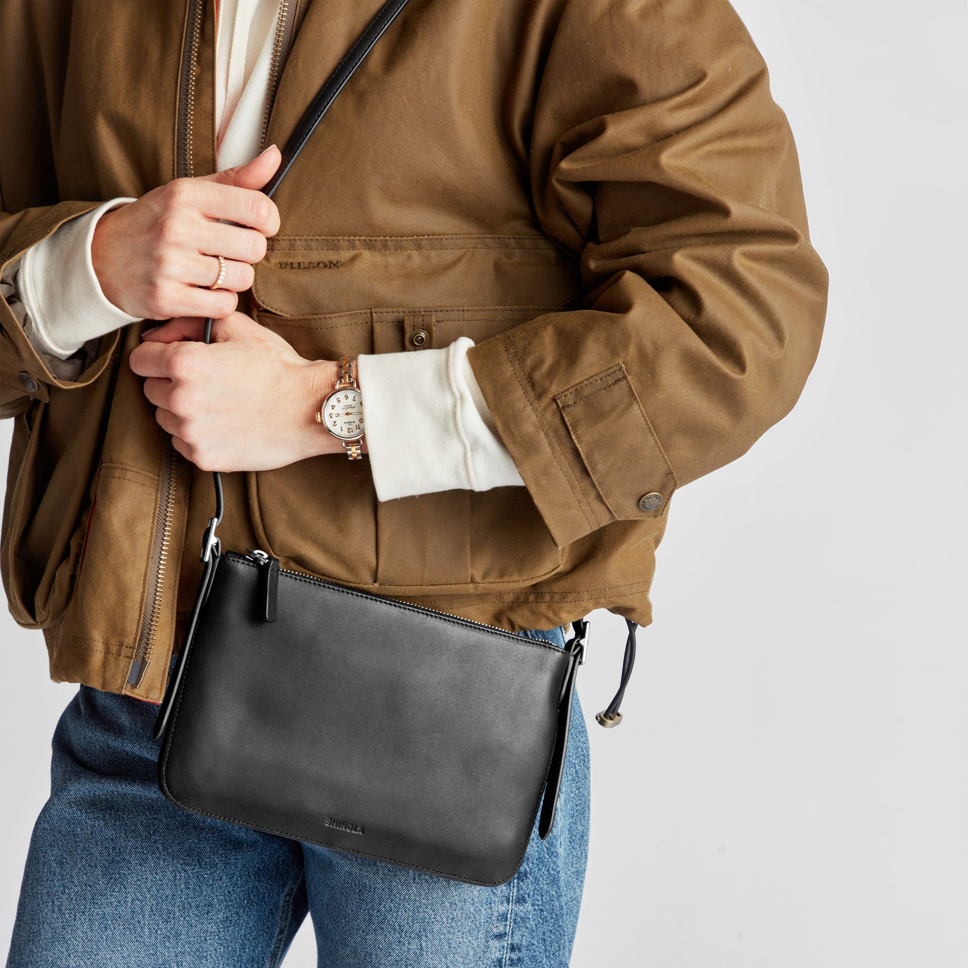 A person in a brown jacket and blue jeans holds the Shinola Small Bixby Crossbody - Black and wears a light-strap wristwatch. Their face is not visible.