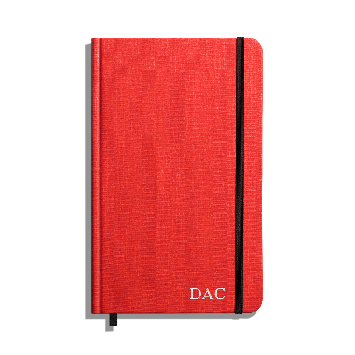 The Shinola Medium Hard Ruled Journal - Tomato is a red hardcover notebook with acid-free paper, black elastic band, black ribbon bookmark, and white DAC initials on the bottom right corner of the cover.