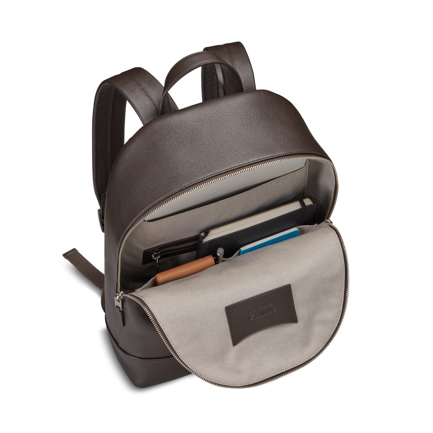The Canfield Backpack - Espresso by Shinola features a brown leather design with an open zipper revealing a spacious interior pocket, room for a notebook and laptop, pen slots, and a beige fabric lining with a stitched label on the inside flap.