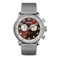 The Shinola Canfield C56 Chronograph Watch - Whiskey features a silver stainless steel mesh bracelet, brown dial with white subdials, silver hour markers, blue and orange accents, and two side pushers for a sleek mid-century modern look.