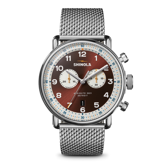 The Shinola Canfield C56 Chronograph Watch - Whiskey features a silver stainless steel mesh bracelet, brown dial with white subdials, silver hour markers, blue and orange accents, and two side pushers for a sleek mid-century modern look.