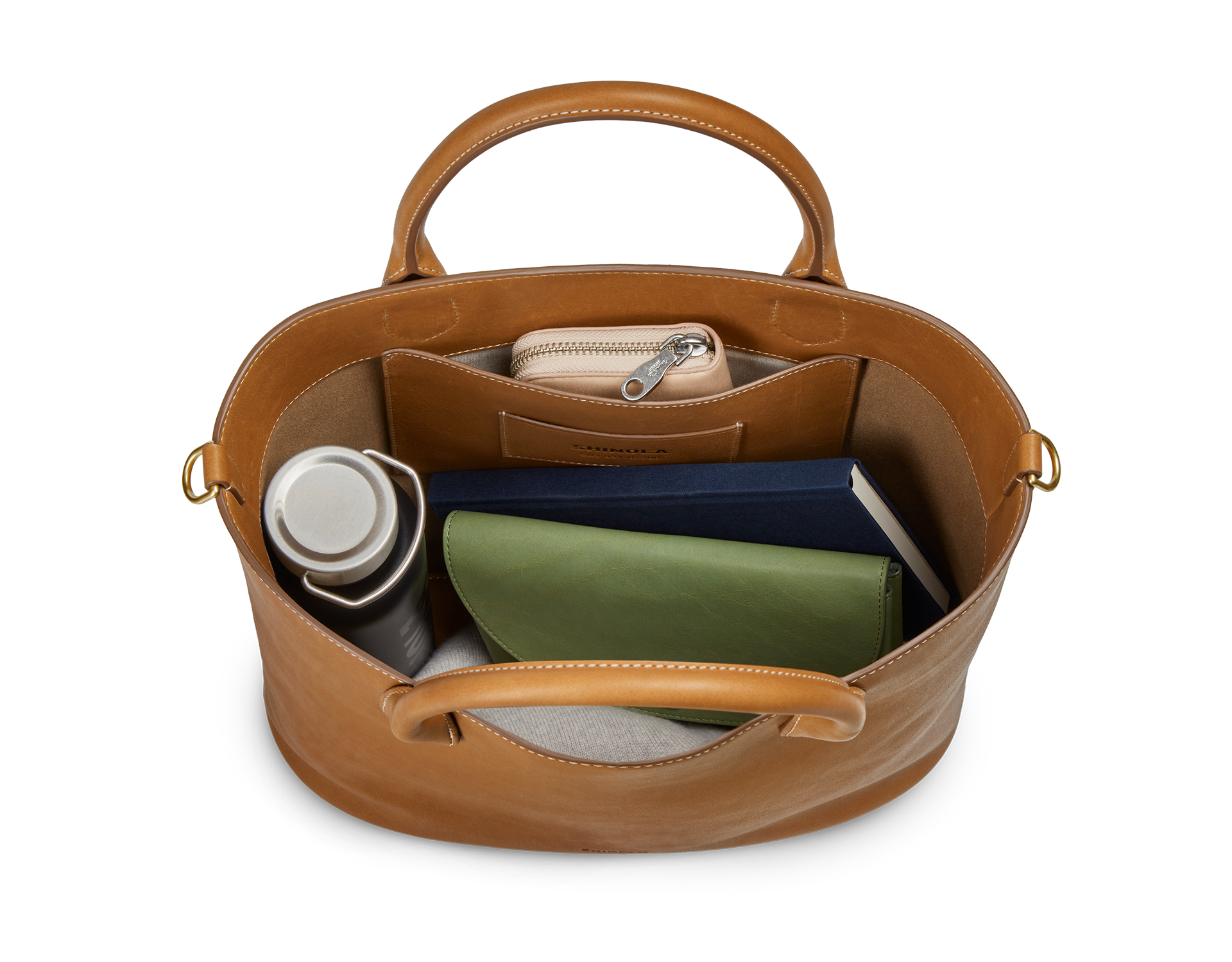 Top view of the Shinola 12 Hour Tote Bag in tan, displaying its roomy interior pocket neatly storing a silver water bottle, green wallet, navy notebook, and beige pouch—ideal for carrying your daily essentials.