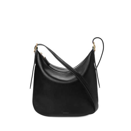 The Shinola Medium Bixby Crossbody - Black features a curved, slouchy shape in natural leather with gold-tone hardware and two adjustable straps, displayed against a plain white background.