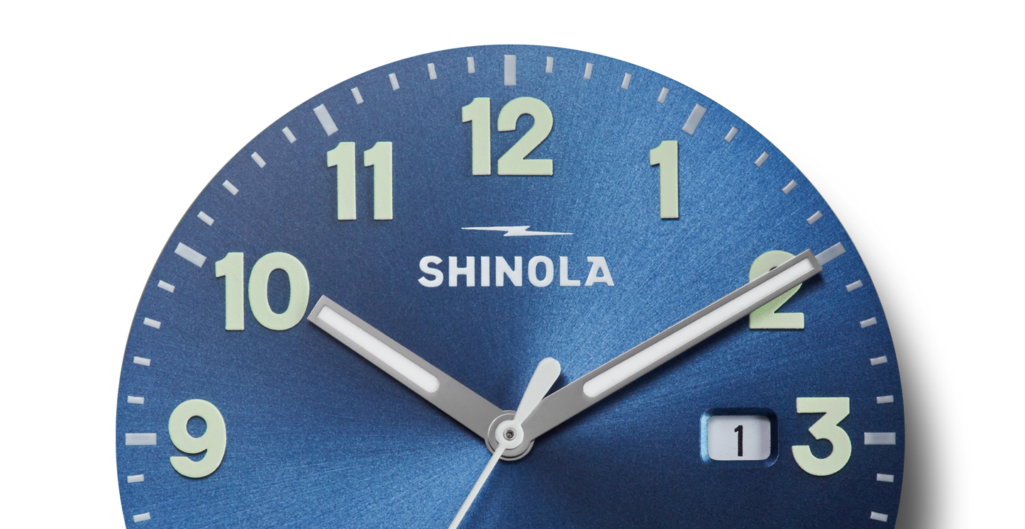 Close-up of a Shinola Detrola Watch - Denim Blue shows the time as 2:08. The watch features large, luminous numbers and hands, and a date window showing "1" next to the 3 o’clock position.