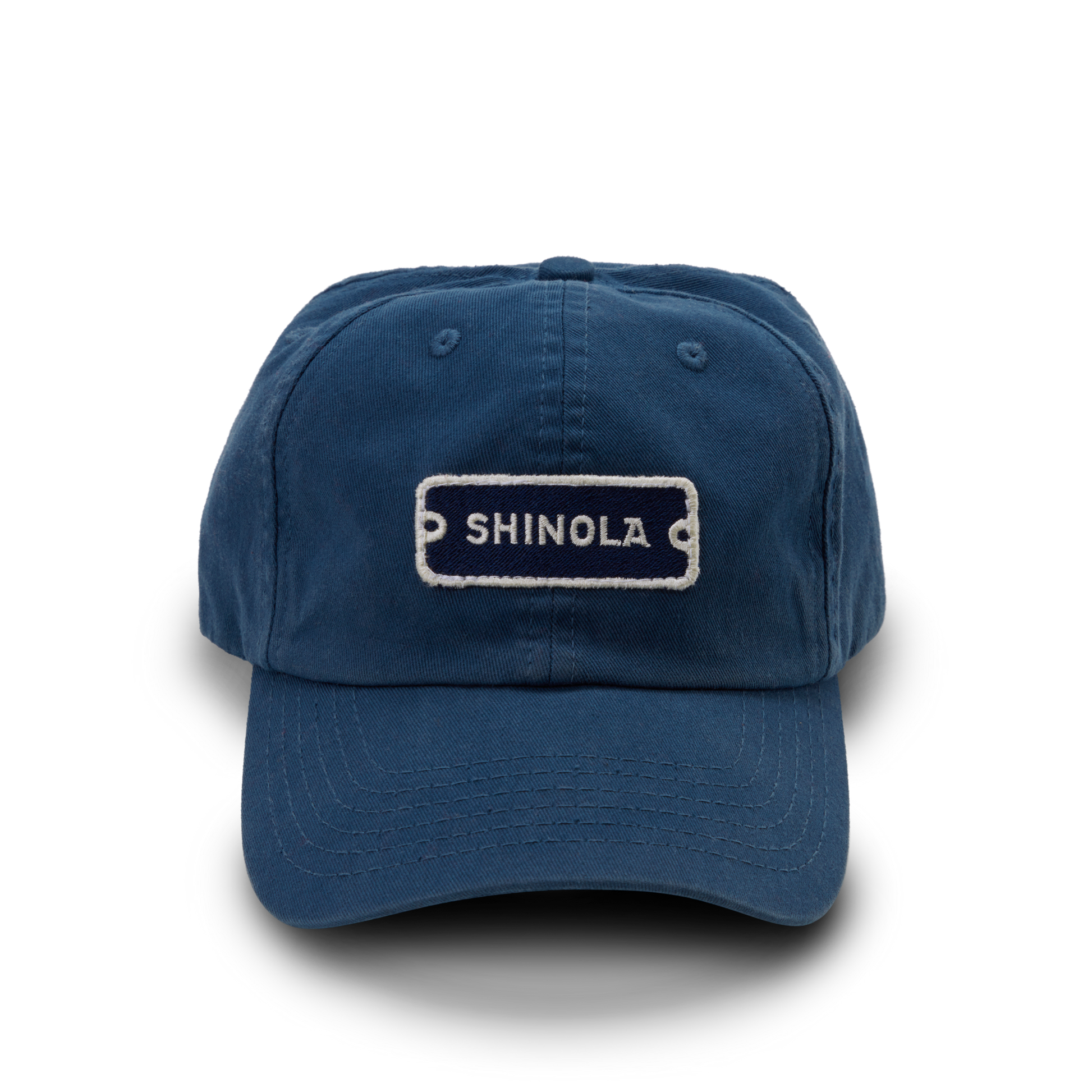 The Shinola Cotton Twill 6 Panel Cap is crafted from garment-dyed cotton twill in blue, featuring a white-stitched SHINOLA patch, visible stitching, curved brim, and a leather strap closure.
