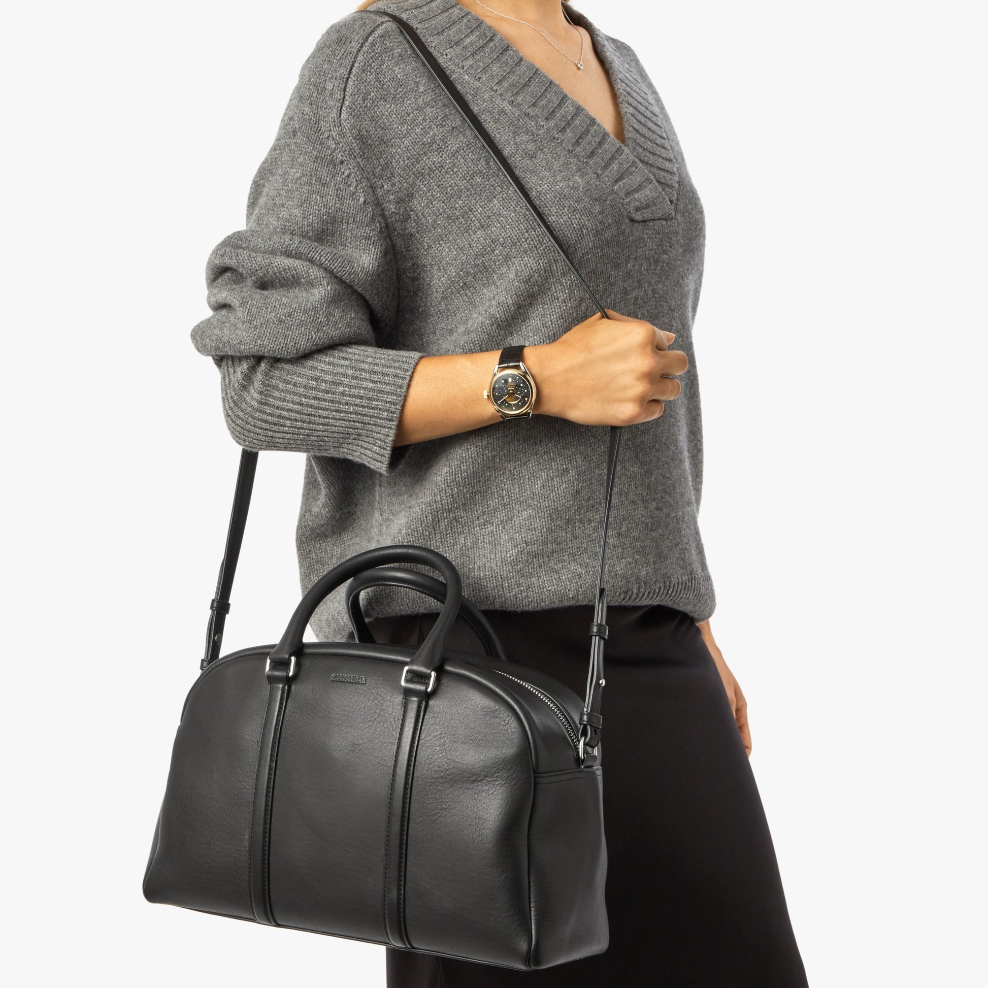 A woman in a gray sweater and black skirt carries the Shinola Runwell Satchel - Black, a large leather shoulder bag with two handles and a strap. She completes her look with a black wristwatch and a necklace.