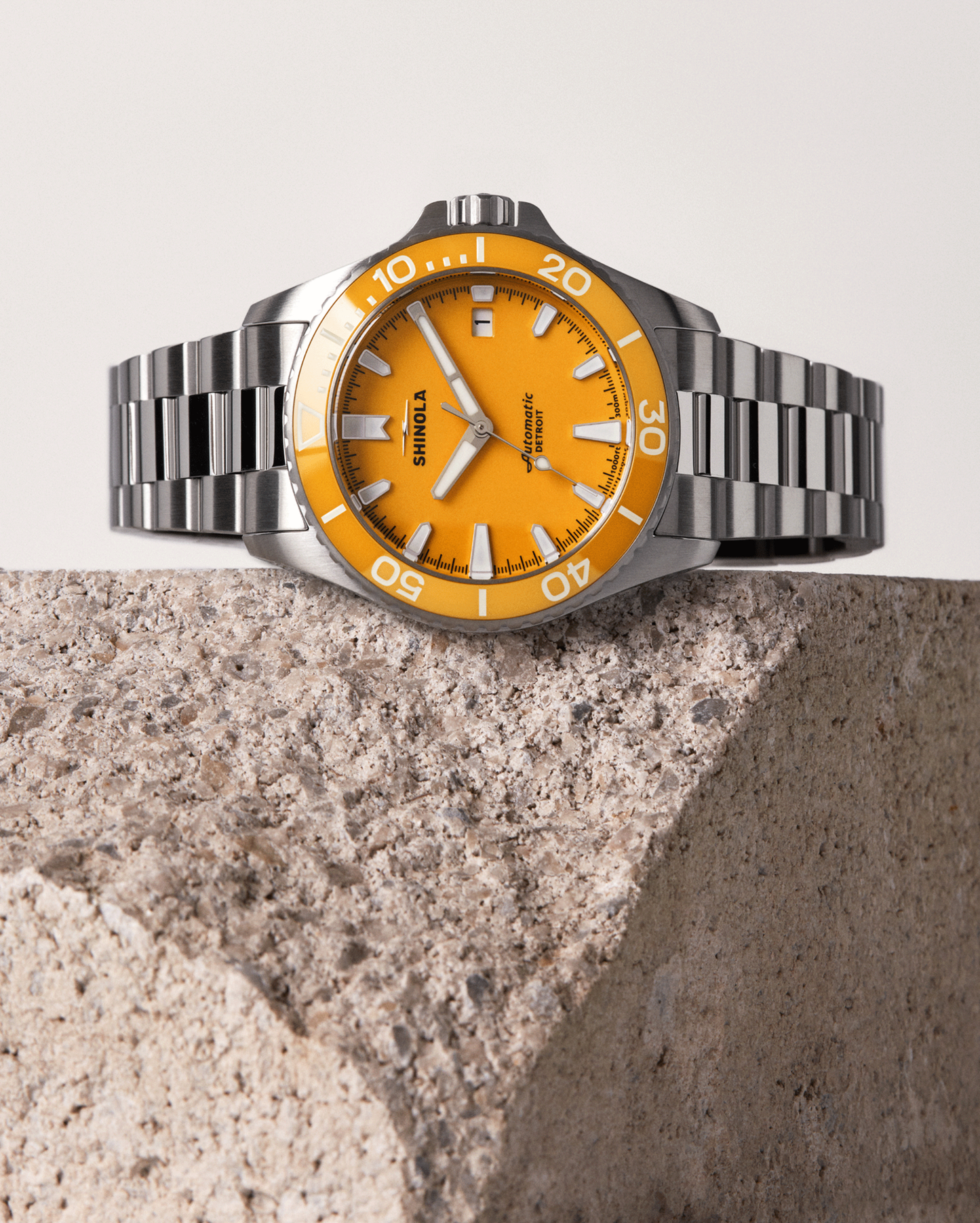 A stainless steel Shinola Lake Huron Monster Automatic Dive Watch with a vivid orange dial and silver hour markers sits on a textured stone block, set against a neutral backdrop—an eye-catching accessory for any occasion.