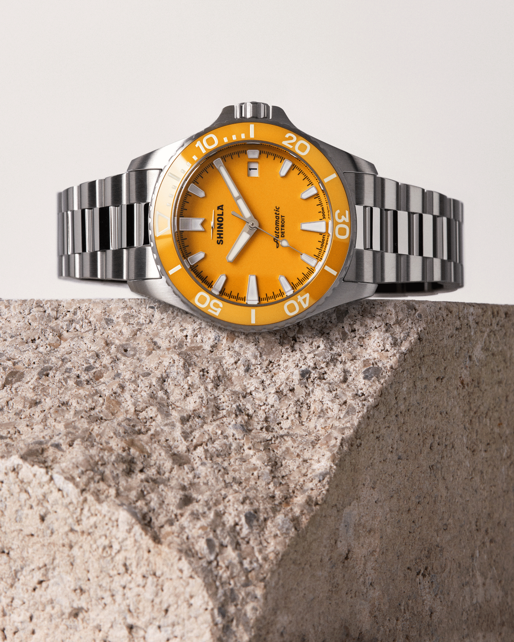 A stainless steel Shinola Lake Huron Monster Automatic Dive Watch with a vivid orange dial and silver hour markers sits on a textured stone block, set against a neutral backdrop—an eye-catching accessory for any occasion.