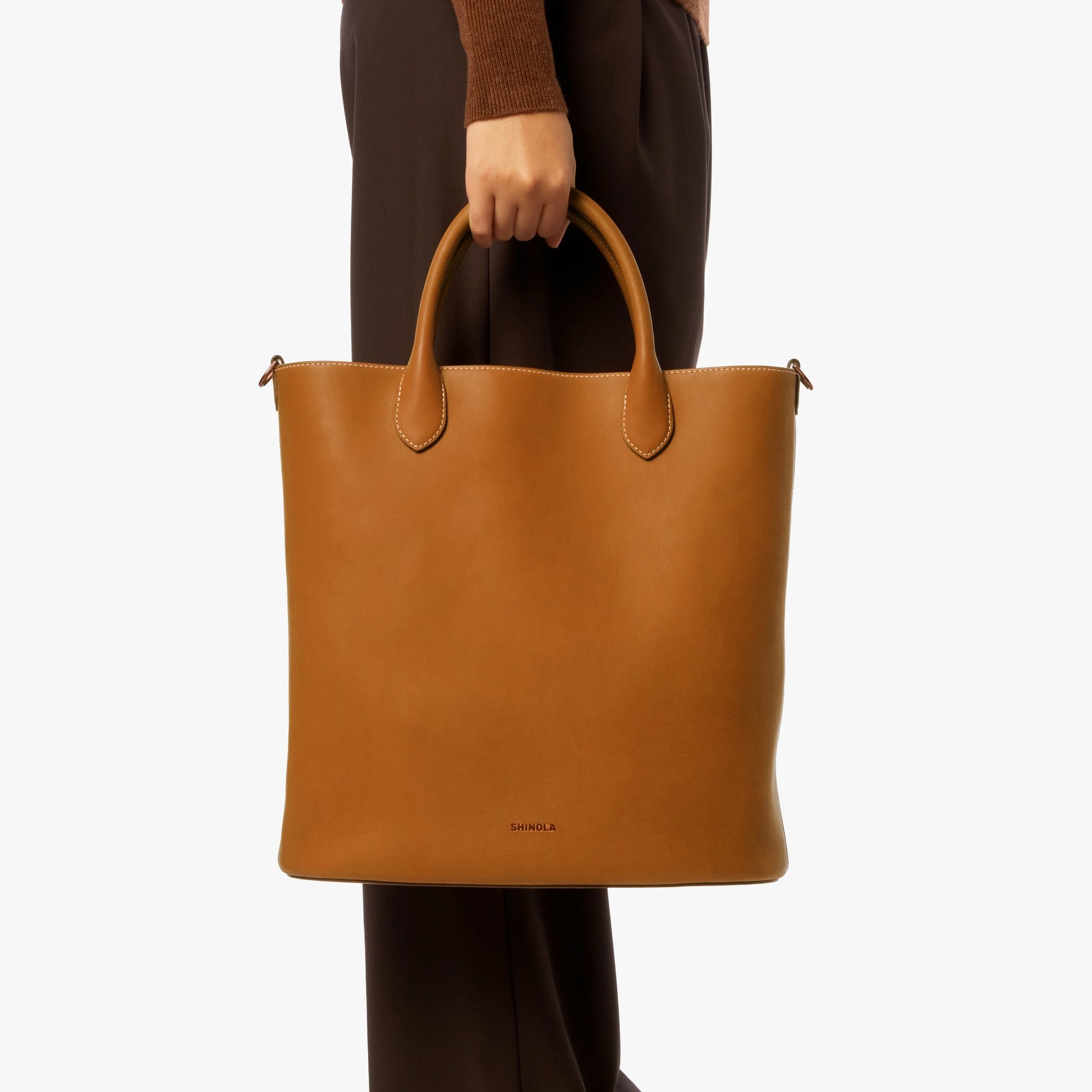 A person in dark brown pants and a brown sweater holds the Shinola 12 Hour Tote Bag in Tan—a spacious, stylish leather tote with two handles—ideal for daily essentials, shown against a plain white background.