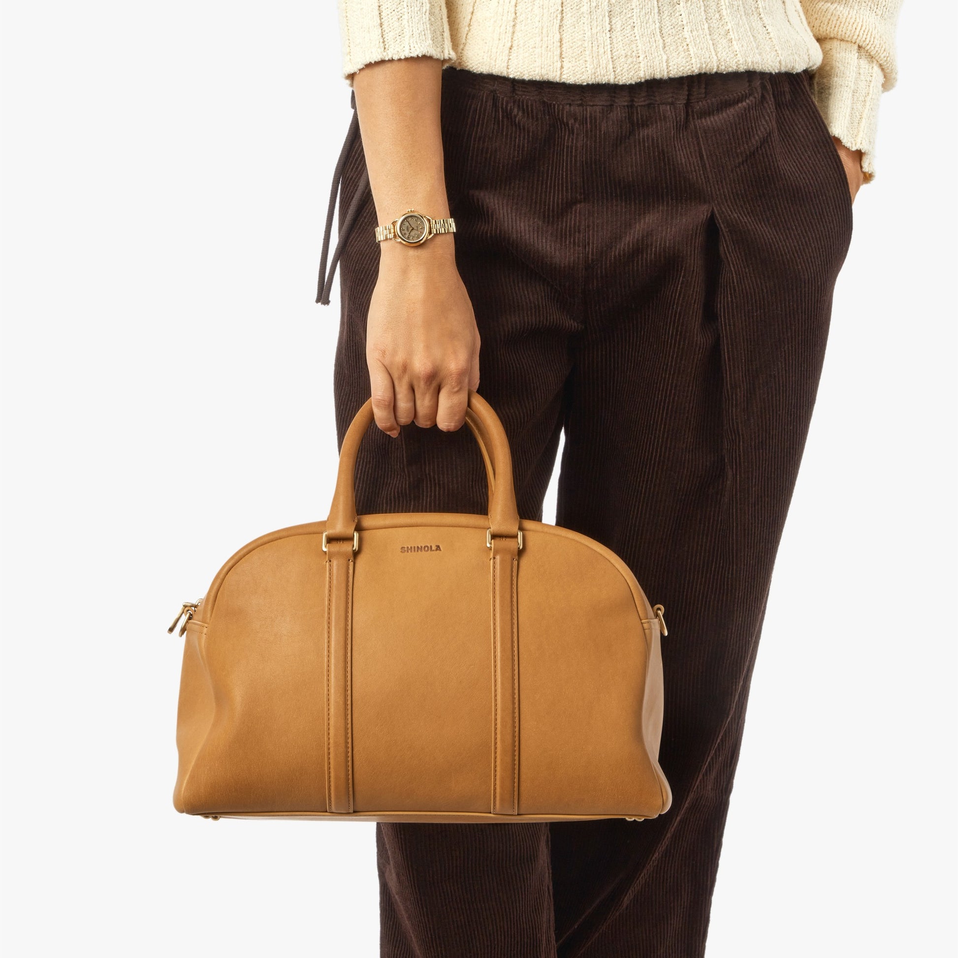 A person in a cream sweater and brown pants holds the Shinola Runwell Satchel - Honey, crafted from premium natural leather, by the handles. They wear a watch on their left wrist. The background is plain white.